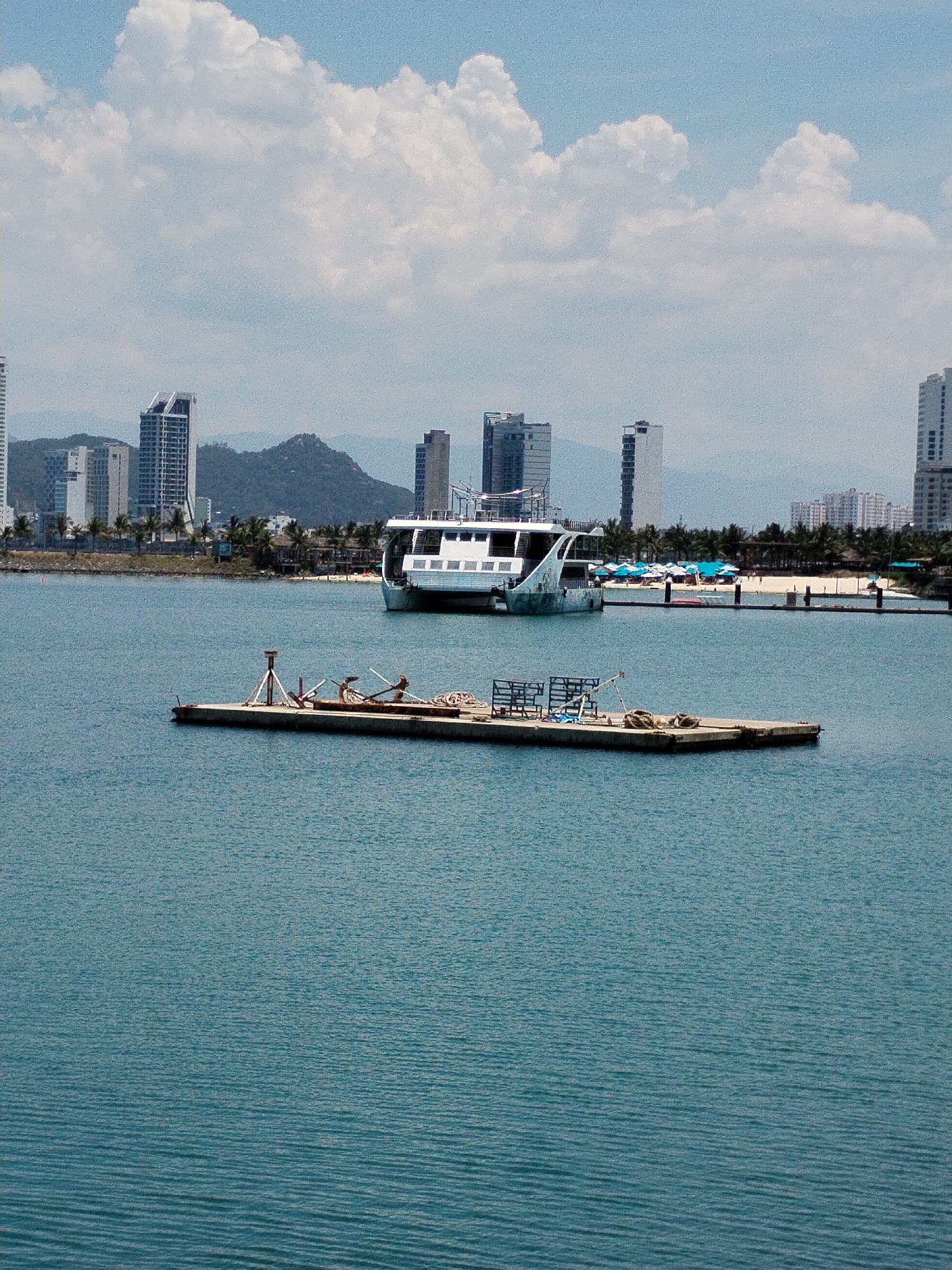 The image depicts a waterfront scene in Bac Nha Trang Ward, Vietnam. In the foreground is a concrete platform with a metal tripod and what appear to be discarded boat anchors and other equipment. Further out in the water is a modern, white ferry. The shoreline is lined with palm trees and buildings, including tall apartment towers. A beach with blue umbrellas and people can also be seen. In the background, rolling green hills blend with a partly cloudy sky.