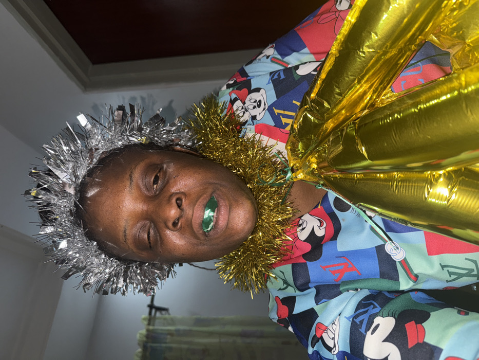 A single individual is depicted in a close-up, chest-up shot, facing the camera directly in an indoor setting located in Sabon Tasha, Nigeria. The person has dark skin; their right eye (viewer's left) is partially closed, and their left eye (viewer's right) is squinted. A green, oval-shaped object is visible between their lips. They are wearing a silver, spiky tinsel-like headpiece on their head and a golden, fluffy tinsel-like garland around their neck. The individual is dressed in a long-sleeved garment patterned with various cartoon characters, specifically Mickey Mouse and Minnie Mouse, alongside what appear to be luxury brand logos (e.g., Louis Vuitton, Gucci) on a multi-colored background primarily featuring shades of light blue, red, green, and black. They are holding a cluster of approximately three to four elongated, shiny, gold-colored balloon-like objects, secured with a thin green string that is tied under the golden neck garland. The background is blurred, showing a light-colored curtain on the left and a darker, indistinct area to the right. The scene is illuminated by a direct light source.