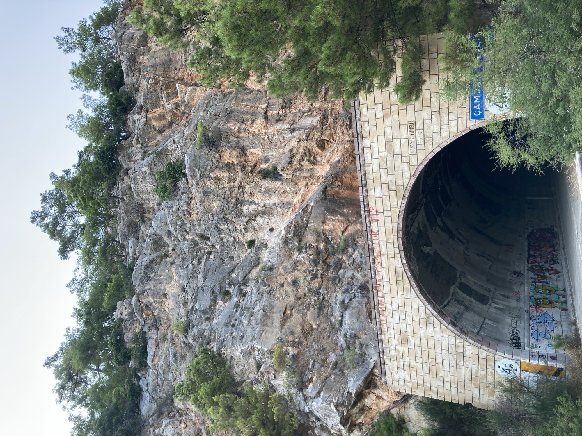 The image shows the entrance to a tunnel named Çamdağ Tüneli in Kemer, Turkey. The tunnel is carved into a rocky hillside covered with green trees. The entrance is framed by light-colored brickwork, and the interior of the tunnel is dark and decorated with graffiti. A road leads into the tunnel. There are also road signs visible, one indicating a height restriction of 4 meters. The overall scene suggests a natural landscape with man-made infrastructure.
