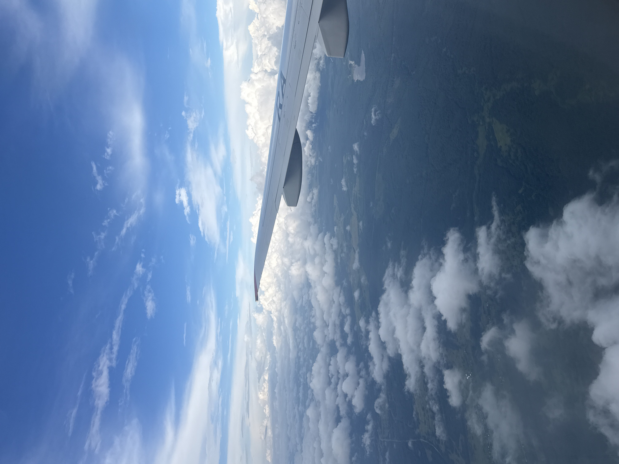 An aerial view from an airplane window shows a vast expanse of clouds and a green, forested landscape below. The airplane's wing is visible in the foreground, extending into the right side of the frame. Scattered white clouds drift across a bright blue sky dotted with wispy cirrus clouds. Below the clouds, a dense, dark green forest covers the land, with occasional clearings and a small body of water visible. The image appears to capture the Russian countryside, potentially near Staroe Mart'yanovo, given the context. No people or specific objects are discernible in the landscape, emphasizing the natural environment.