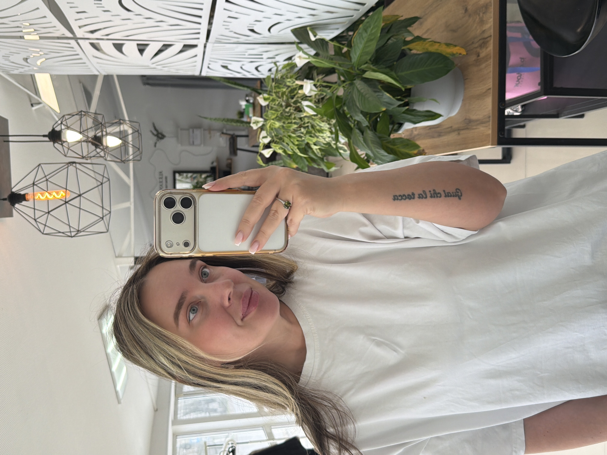 A young woman with light brown hair highlighted with blonde streaks and blue eyes is captured in a mirror selfie. She wears a plain white t-shirt and displays a French manicure on her nails. A ring with a green stone adorns her right ring finger, and a tattoo reading "qual chi la tocca" is visible on her inner right forearm. She is holding a gold-cased smartphone, likely a recent iPhone model, which she uses to take the photo.
The reflection reveals a modern indoor setting. The upper background features white decorative ceiling panels with intricate geometric cutouts. Below these, three black metal cage-style pendant lights with exposed Edison bulbs hang, two of which are illuminated. A wooden table or counter stretches across the middle-right of the frame, supporting two potted plants: a larger one with dark green leaves and white blossoms, and a smaller, variegated plant. A black metal shelving unit is partially visible on the far right. A window reflecting outdoor light is seen in the lower left, hinting at a daytime setting. The overall atmosphere is bright and contemporary. The location context for this scene is Chaikovsky, Russia.