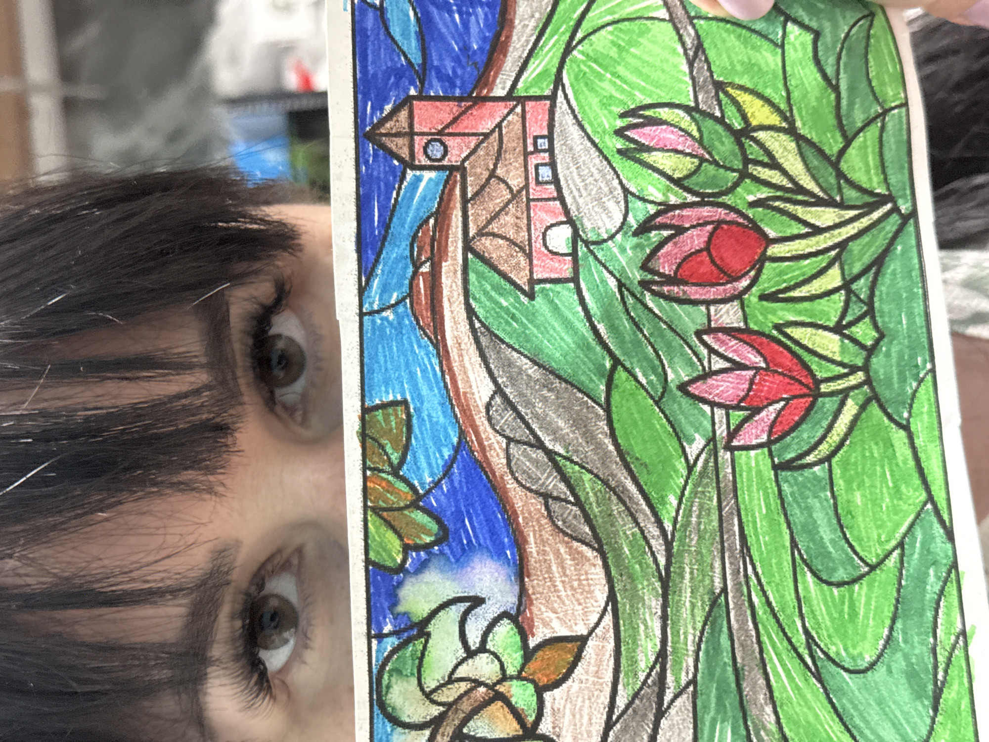 A person with dark hair and brown eyes is holding a colored drawing. The drawing is a stained glass style image featuring a house, flowers, leaves, and water. The colors are primarily red, pink, green, and blue. The location in Zav'yalovo, Russia is not discernible from the image.