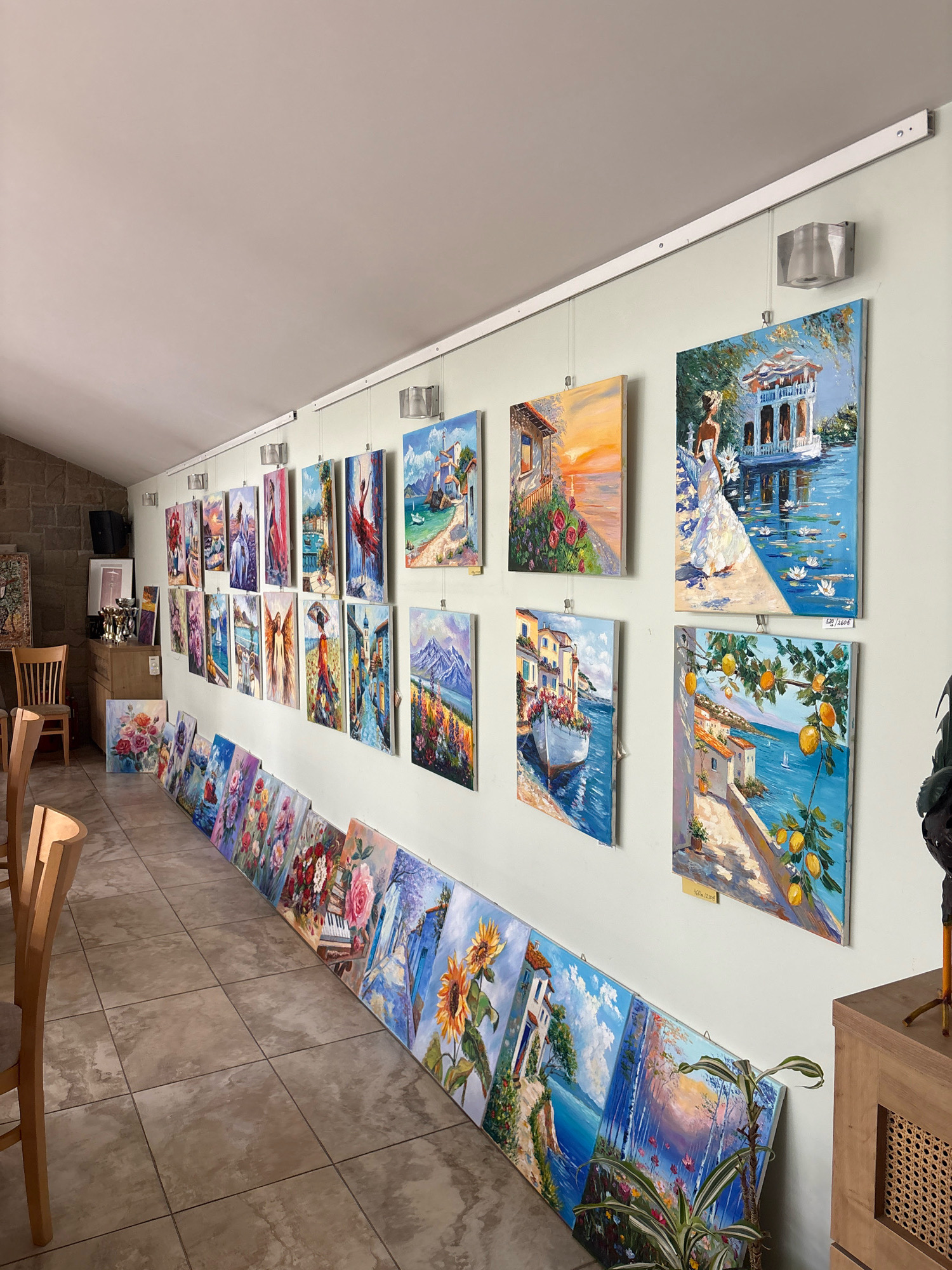 The image depicts an art gallery, potentially located in Ustina, Bulgaria, exhibiting numerous oil paintings. The artworks are displayed both on the wall and leaning against it on the floor. The paintings feature a variety of subjects, including landscapes, seascapes, floral arrangements, and figurative art.
On the wall, paintings are hung from a track system, illuminated by track lighting. These include scenes of coastal towns with boats, a woman in a white dress by a body of water, a sunset over a building, and a mountain landscape. Further along the wall, more paintings show vibrant colors and detailed brushwork.
Leaning against the wall on the floor is a collection of canvases, predominantly featuring floral motifs and serene landscapes. These include depictions of roses, sunflowers, and other flowers, as well as idyllic scenes of houses by the sea and forests.
The environment suggests a dedicated space for art display. The floor is tiled with neutral-colored tiles. Wooden chairs with cushioned seats are placed in the foreground, indicating seating for visitors. A stone wall is visible on the left side of the image, adding to the rustic ambiance. To the right, a wooden cabinet with woven details and a decorative bird sculpture are present. The overall impression is a well-curated exhibition showcasing a diverse range of artistic talent.