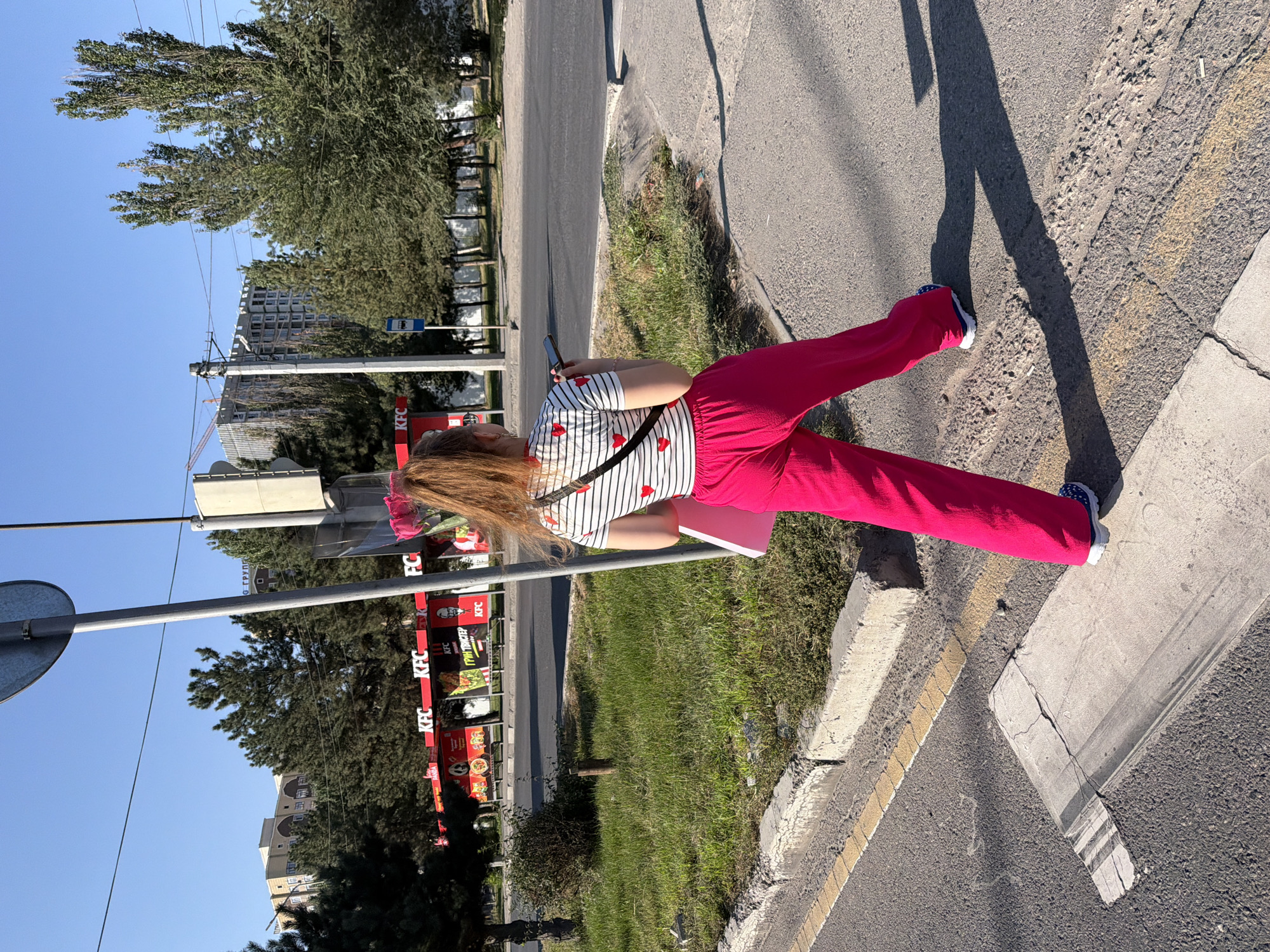 In this full-shot, eye-level, outdoor photograph, a young woman with auburn hair pulled back in a high ponytail walks away from the camera on a sunny day in an urban environment. She is wearing a striped t-shirt with small red hearts, bright pink wide-leg pants, and blue and white sneakers. A black crossbody bag is slung over her shoulder. She holds a dark-colored smartphone in her right hand. A light pink flat object, possibly a folder or book, is held in her left hand. The woman is walking on a paved sidewalk next to a street. Across the street, a KFC restaurant with red signage and outdoor seating is visible, along with trees, buildings, and a construction site in the background. The ground in the foreground is asphalt with a crosswalk marked by white paint. The lighting suggests it is late afternoon, casting long shadows.