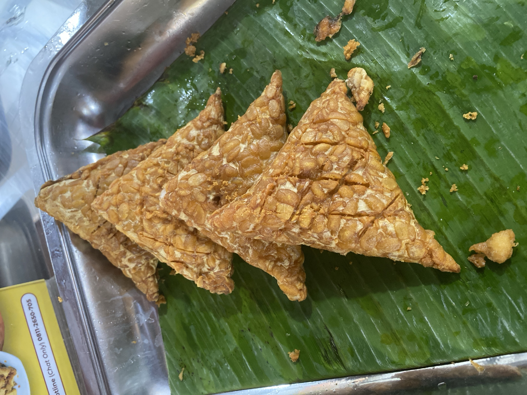 This media file shows a close-up, overhead view of several triangular pieces of fried tempeh arranged on a bright green banana leaf. The tempeh is golden brown and appears crispy, with visible lines indicating it was cut into shape. Small, scattered pieces of fried batter or tempeh are visible on the banana leaf around the main pieces. The banana leaf itself has a glossy sheen, suggesting it might be slightly wet. The tempeh is placed on a stainless steel tray, which takes up the left side of the frame. Part of a yellow sticker with black text is visible on the edge of the tray. The text is partially obscured but includes "Hotline (Chat Only) 0811-2650-705". The scene appears to be a food stall or market setting, likely in North Jakarta, Indonesia, given the visual cues of common food presentation and the style of the text. The lighting is bright and appears to be artificial, possibly from overhead lights in an indoor setting or during the day. There are no people or animals visible in the frame. The focus is solely on the food presentation.