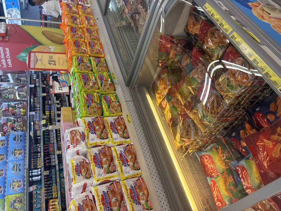 A supermarket aisle displays instant noodles and frozen food in a refrigerated section. Various flavors of "Mie Sedaap" instant noodles are stacked on shelves, including "Goreng" (fried) variants in orange, green, and white packaging. Frozen food items, such as potato products, are visible behind a glass door. Price tags and promotional signs advertise discounts on the products. The layout showcases a typical grocery store setup with categorized items arranged for easy browsing and purchase.