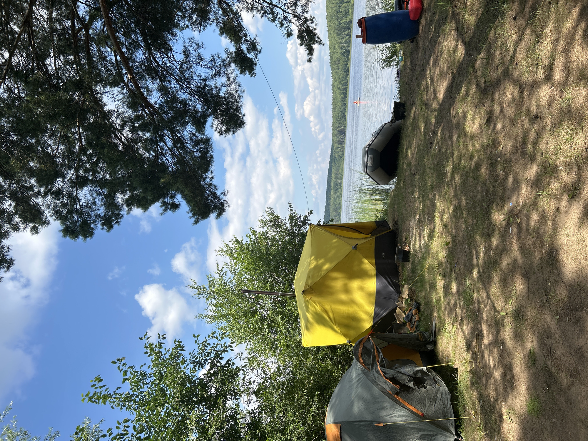 A campsite is set up on the sandy, grassy shore of a large body of water, likely a lake or river, in a wooded area. Two tents are pitched: one is a gray and orange dome tent, and the other is a larger, yellow cube-shaped tent with a visible chimney pipe. In front of the tents, there are logs and some camping gear. An inflatable boat is resting on the bank near the water's edge. To the right, a large blue barrel and a red container are visible. In the distance, across the water, a treed shoreline is visible, and a person can be seen on a paddleboard. The sky is blue with scattered white clouds, and sunlight filters through the branches of a pine tree in the foreground. The location is identified as the city of Chaykovsky, Russia.