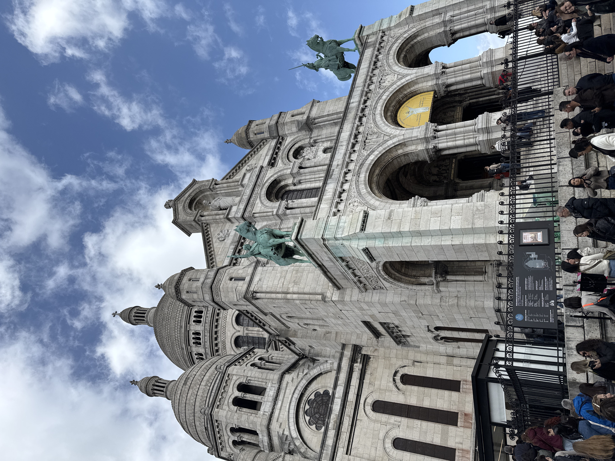 The image captures a portion of the Sacré-Cœur Basilica in Paris, France, viewed from below at an oblique angle. The building's architecture, characterized by white stone and multiple domes, is prominently displayed against a backdrop of blue sky with scattered clouds. Two bronze equestrian statues adorn the basilica's facade. A crowd of people is present behind a black metal railing, some are observing the building and potentially reading a nearby information panel, and others are looking in different directions. The image is taken at daytime.