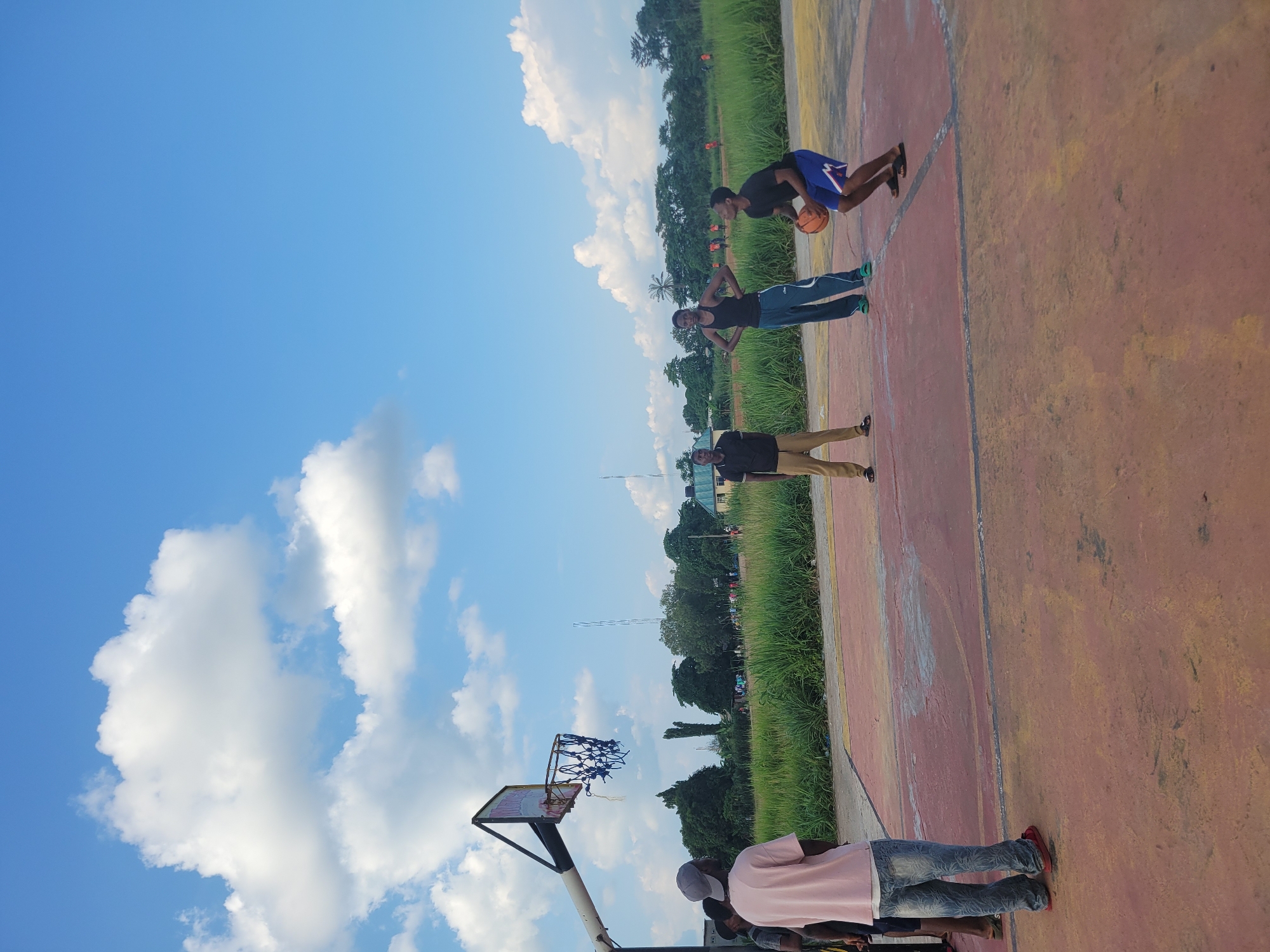The image shows four people on an outdoor basketball court. The court is made of a reddish-orange material, with yellow and white markings. There is a basketball hoop and backboard on the left side of the frame. Tall green grass borders the court, and beyond that, trees and some buildings are visible. The sky is a bright blue with scattered white clouds, suggesting it is daytime and the weather is pleasant.
One person, wearing a black t-shirt and blue shorts, is in mid-action, holding a basketball and appearing to dribble or prepare to shoot. Another person, in a black tank top and blue pants, stands with hands on hips. A third person, in a black shirt and khaki pants, stands further down the court. The fourth person, wearing a light pink shirt and jeans, is partially visible in the foreground. Their poses suggest they are engaged in a casual game or practice session. The overall mood appears relaxed and active, typical of an outdoor recreational setting. No visible text is present in the image. The context provided indicates the location is Egbagu, Nigeria.
