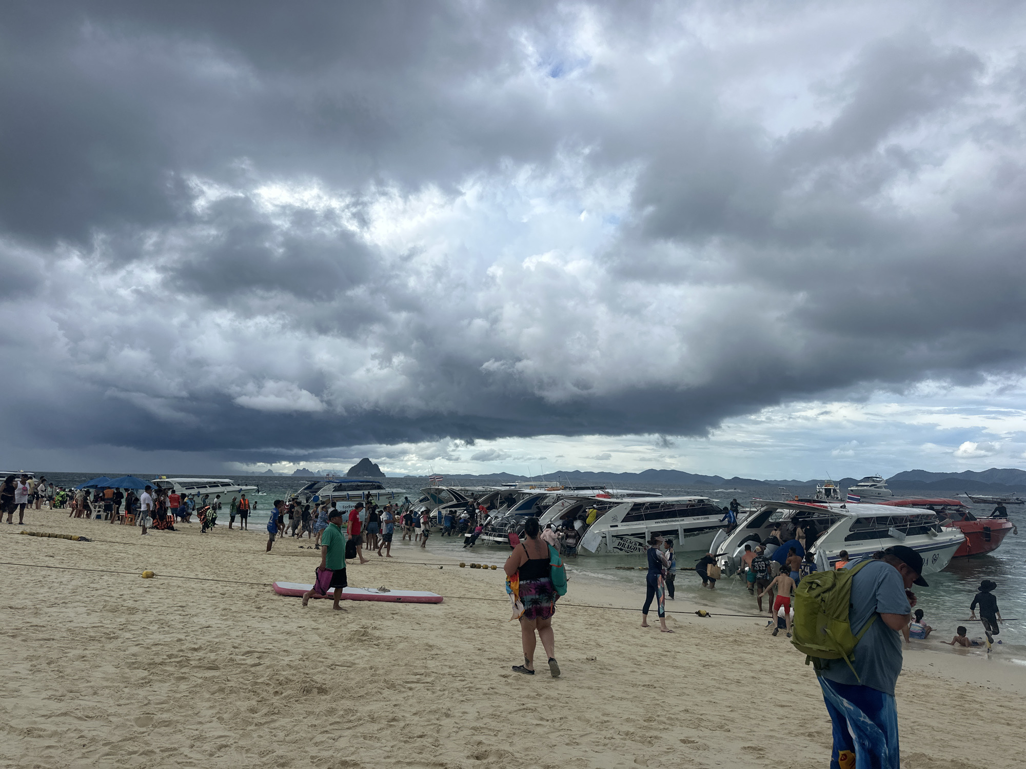 A crowd of people are gathered on a sandy beach in Tambon Phru Nai, Thailand, under a heavily overcast sky. Numerous speedboats are moored in the shallow water near the shore, with some people boarding or disembarking from them. The beach is lined with more speedboats, and in the background, distant islands are visible through the hazy atmosphere. People are dressed in casual beach attire, and some are carrying bags or wearing backpacks. The overall scene suggests a popular tourist destination with active boat tours.