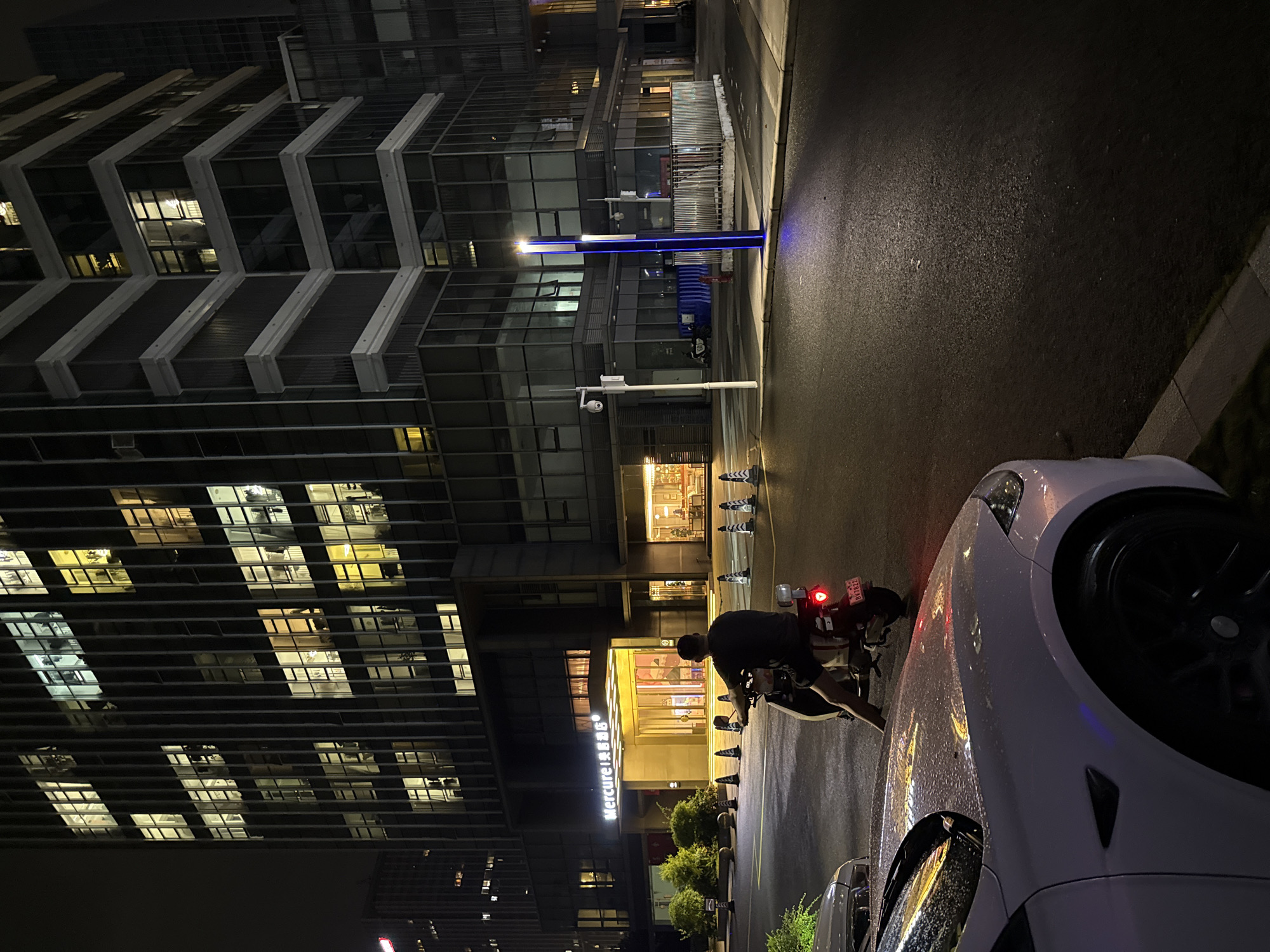 An exterior, ground-level, medium shot captures a nighttime scene in the Huangpu District, China, outside the Mercure hotel. A person, seen from behind, is seated on a scooter. The scooter is parked on a wet, dark asphalt road, with a white car to the left also appearing wet and reflecting the building's lights. In the background, a multi-story building displays numerous illuminated windows, suggesting it is occupied or actively lit from within. A blue illuminated light fixture stands near the center of the frame on the sidewalk. The scene is characterized by artificial lighting from the building and the street lamp.