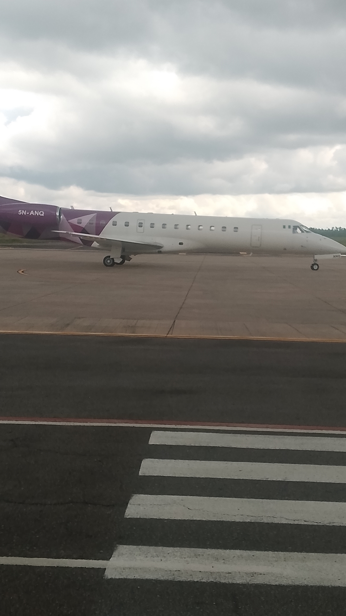 An Embraer ERJ-145LR aircraft, registration 5N-ANQ, is parked on the tarmac at Orerokpe Airport in Orerokpe, Nigeria. The aircraft has a white fuselage with purple accents on the tail and wingtips, featuring a geometric design. The registration number "5N-ANQ" is visible on the rear fuselage. The plane is positioned on a gray asphalt surface, with visible runway markings, including a pedestrian crossing in the foreground. The sky is overcast with gray clouds.