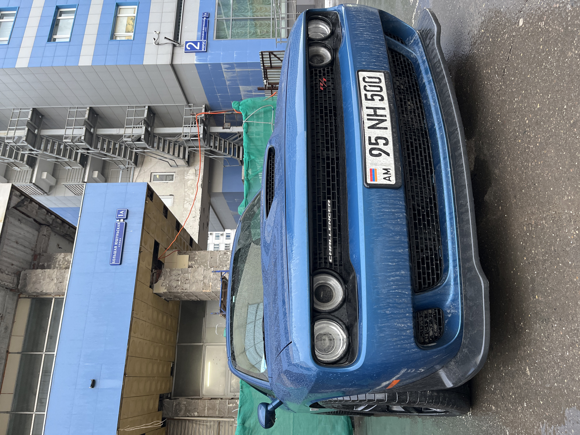 The image shows a blue Dodge Challenger parked on a wet pavement. The car has an Armenian license plate reading "95 NH 500". Buildings are visible in the background. Street signs are visible on the buildings, one reading "Большая Юшуньская улица 1А" and the other reading "корпус 2". Based on the street name in Russian, the location is Moscow, Russia. The weather appears to be rainy or recently rained. The buildings show signs of ongoing construction or renovation.