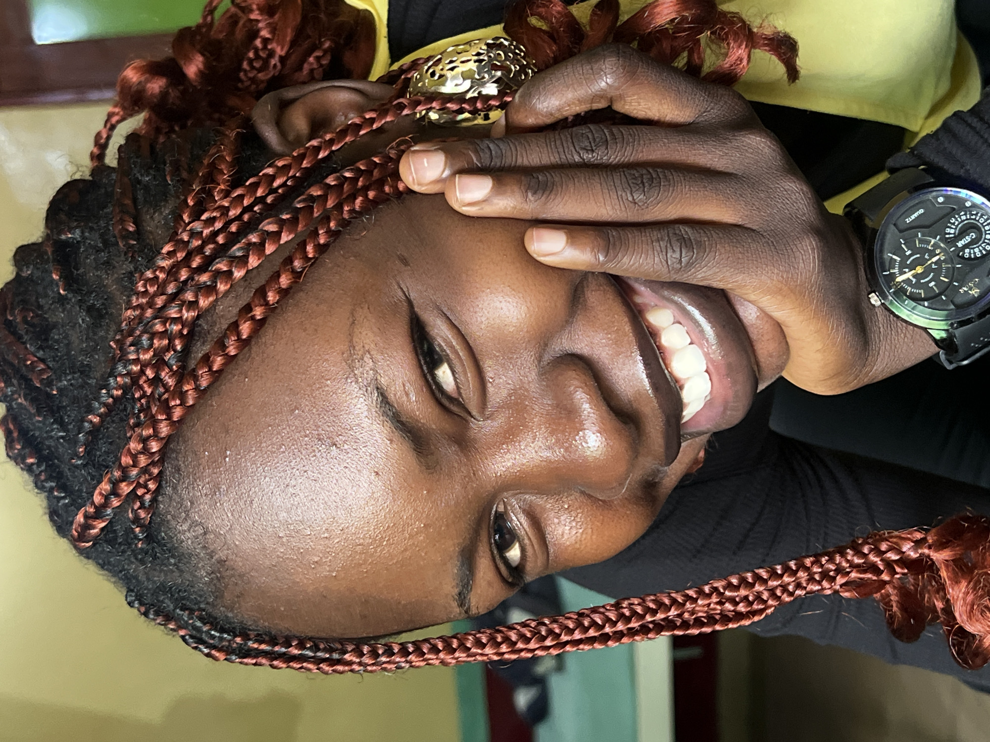 A close-up, eye-level shot shows a young Black woman smiling with her right hand resting on her chin. She has her hair in reddish-brown braids and is wearing a black top and a yellow garment underneath. She also has a watch on her left wrist and a gold earring in her right ear. The background is out of focus, but appears to be an indoor setting with muted colors, possibly in Jos, Nigeria.