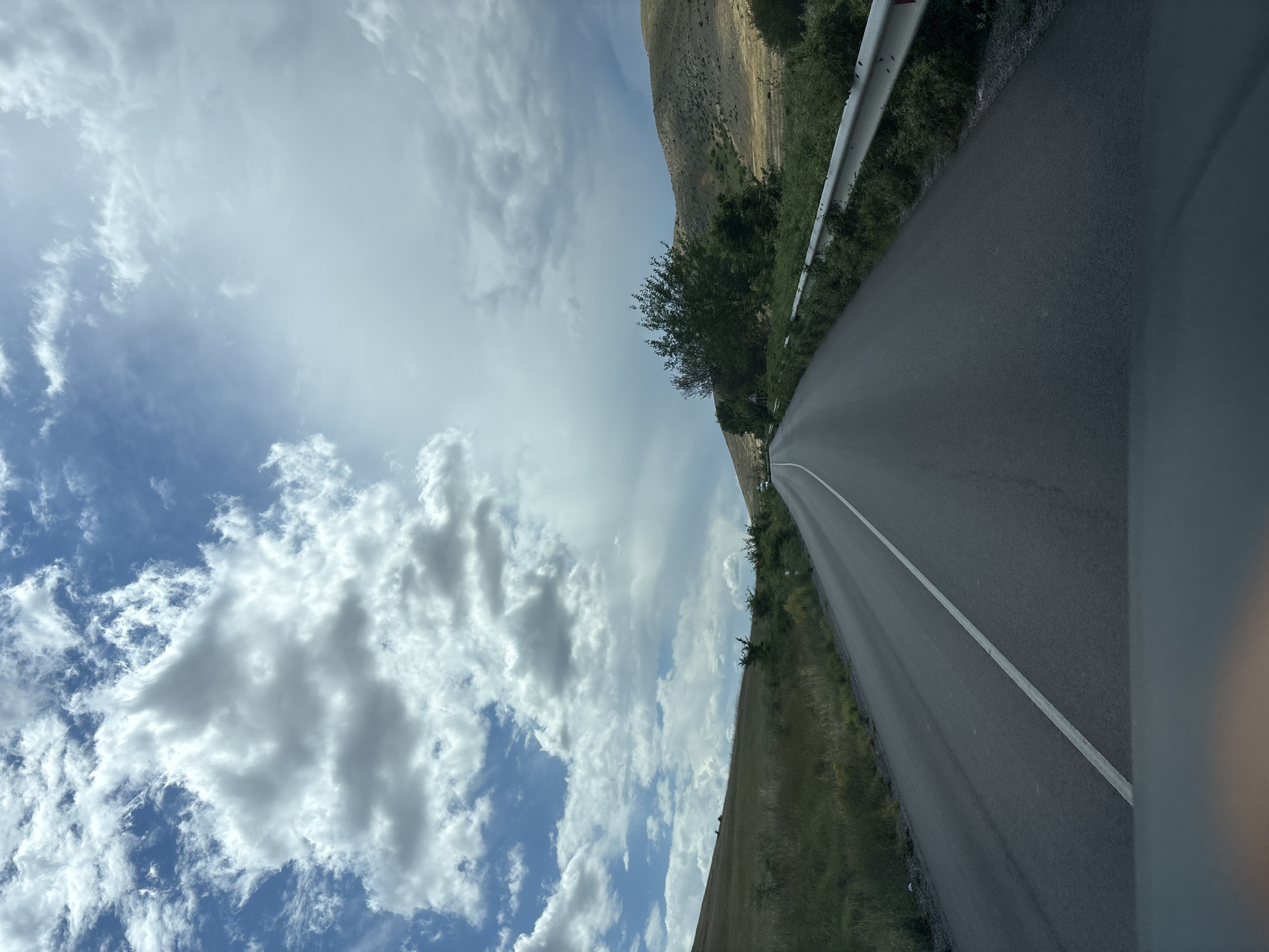 The image depicts a view from a vehicle traveling along a two-lane asphalt road. The road curves slightly to the right, with a white dashed line marking the center. To the left of the road, a grassy hillside slopes upwards, dotted with sparse vegetation. To the right, a metal guardrail separates the road from a dense growth of green bushes and trees. Beyond the guardrail, the terrain consists of a gently sloping, dry, yellowish-brown hillside. In the distance, several trees are visible on the right side of the road, and two white vehicles appear to be on the road ahead. The sky is filled with dramatic, textured clouds, ranging from bright white to darker grays, against a backdrop of blue. The overall impression is that of a scenic drive through a rural landscape under a dynamic sky, possibly in the vicinity of Isperihovo, Bulgaria.
