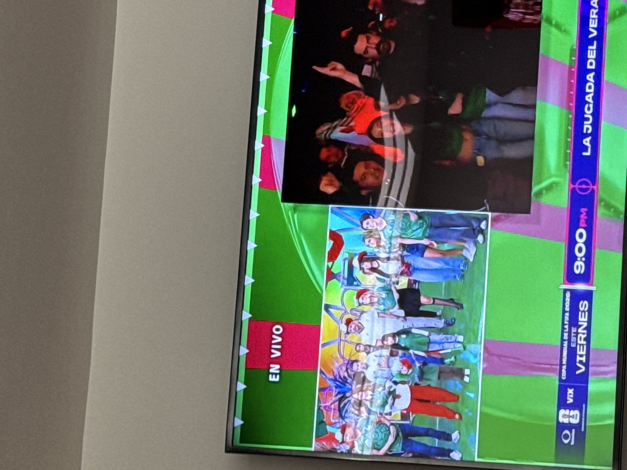 The image is a television screen displaying a live broadcast. The screen is divided into two main sections. The left side shows a group of people, including men, women, and children, standing on a stage with a green and pink backdrop. Many of the individuals are wearing green and white attire, resembling sports fan clothing. The text "EN VIVO" (meaning "live" in Spanish) is prominently displayed at the top of this section.
The right side of the screen is darker and shows a crowd of people with some individuals raising their arms, suggesting an enthusiastic audience.
The bottom portion of the screen contains broadcast information. The text "COPA MUNDIAL DE LA FIFA 2020 ESTE VIERNES 9:00PM LA JUGADA DEL VERANO" is visible. This translates to "FIFA World Cup 2020 This Friday 9:00 PM The Play of the Summer." The presence of "VIX" suggests a media platform or channel.
The image is a captured photograph of the television screen itself, and the television is situated in a room with a plain wall. There is no visual information within the image that allows for the determination of a specific location in San Bernardino Tlaxcalancingo, Mexico.