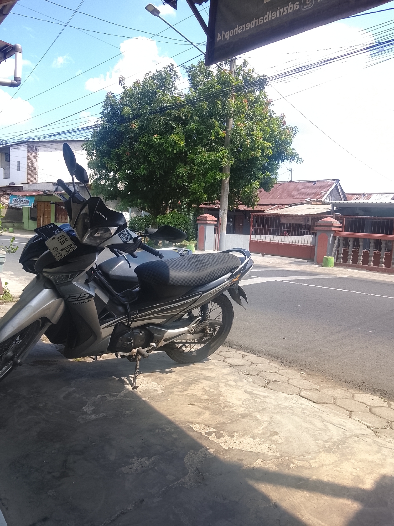 An outdoor, eye-level, medium shot shows a silver Honda motorcycle parked on a concrete sidewalk next to a paved road. The motorcycle is angled towards the left of the frame, with its front wheel pointed slightly outwards. The motorcycle has a black patterned seat and a license plate that reads "R 56 4136 DS 09-27". A tall, leafy green tree stands behind the motorcycle, with its branches extending towards the blue sky. In the background, there are several buildings with red tiled roofs and a red metal fence. Overhead, a network of power lines crisscrosses the sky. A sign with text in a foreign language is visible at the top of the frame. The sun casts shadows on the sidewalk, indicating it is daytime.