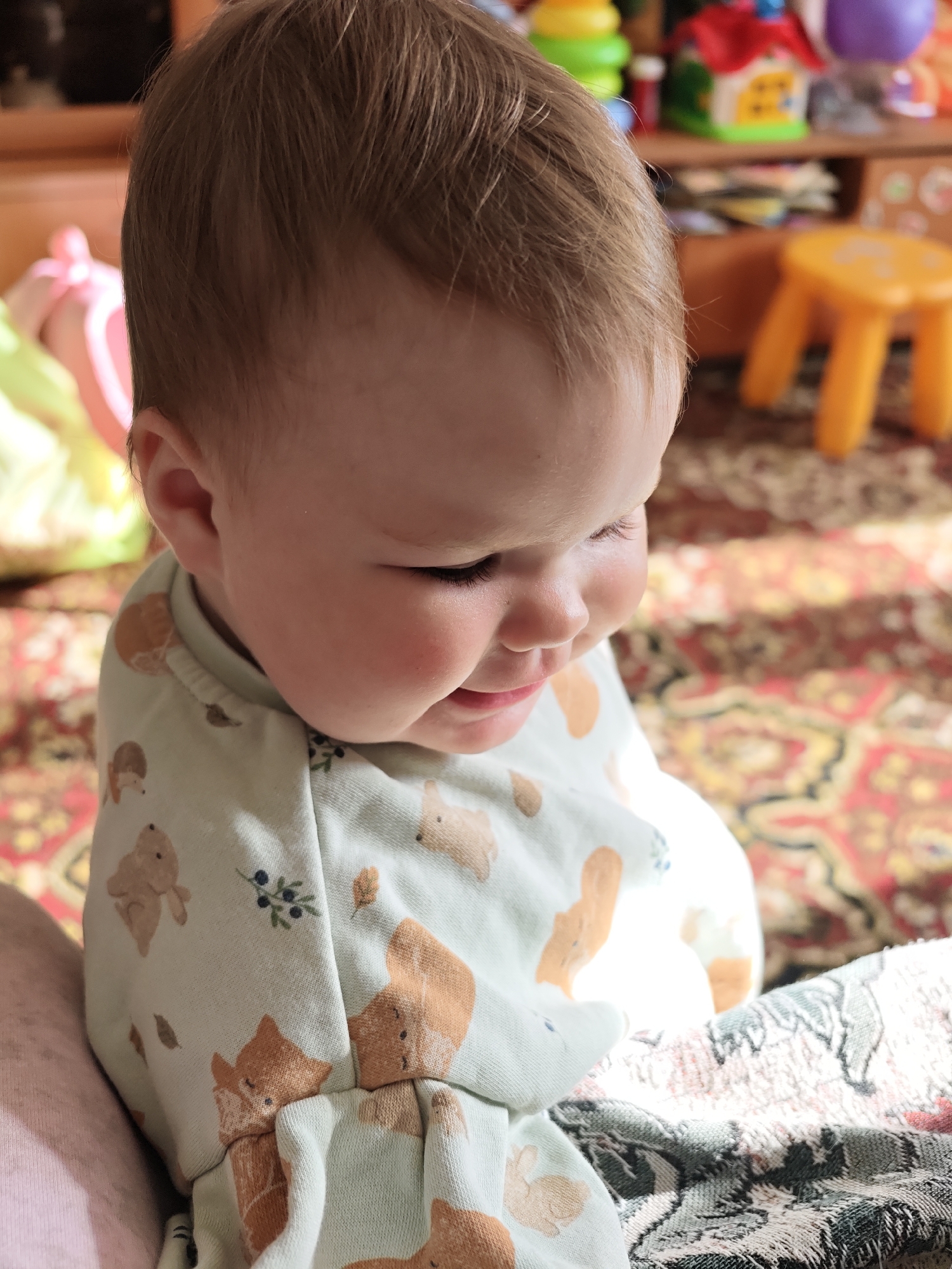 A baby is shown seated indoors, with a light-colored pajama-like outfit featuring prints of foxes, rabbits, and foliage. The baby has light skin and brown hair. The background includes a patterned rug and various toys on a shelf or piece of furniture, including a yellow stool. The setting appears to be a domestic environment, likely a child's room or living area. There is no information to confirm that this image was taken in Уральское, Russia.