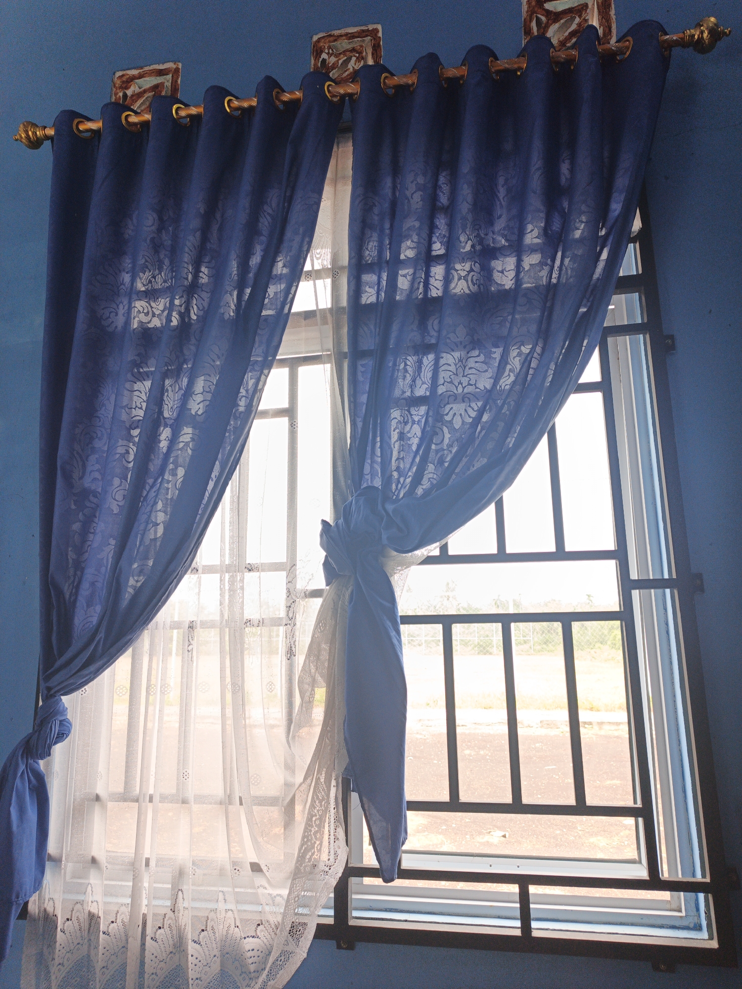 A close-up, eye-level shot captures a window adorned with two layers of curtains. The outer layer consists of thick, dark blue curtains with an embossed floral pattern, draped to the sides. The inner layer features sheer white lace curtains with a delicate, circular pattern, allowing diffused light to pass through. The window itself is framed by a dark metal grate and a white window frame. The wall surrounding the window is painted a vibrant blue. Two decorative square ornaments, with brown and reddish designs, are mounted above the window on the blue wall, affixed to the golden curtain rod. The scene is located inside a room in Resam Lapis, Indonesia.