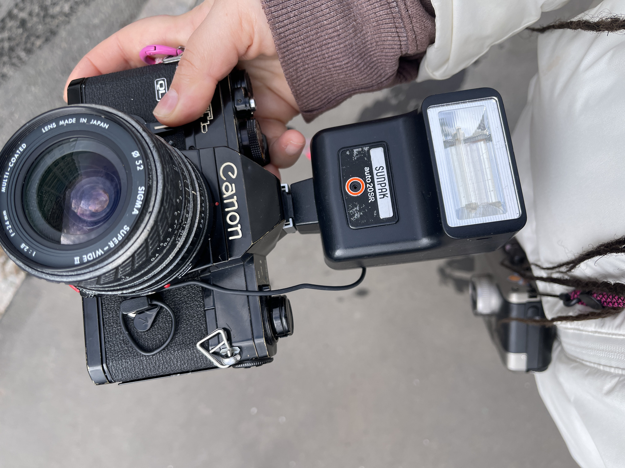 A Canon film camera with a Sigma lens and a Sunpak flash is being held in someone's hand. The lens is marked "Lens Made in Japan, Multi-Coated, Ø 52, Sigma Super-Wide II, 1:2.8 f=24mm." The flash unit is a Sunpak auto20SR. The setting appears to be outdoors, likely in an urban area. There is no definitive information to confirm that this image was taken in Moscow, Russia.