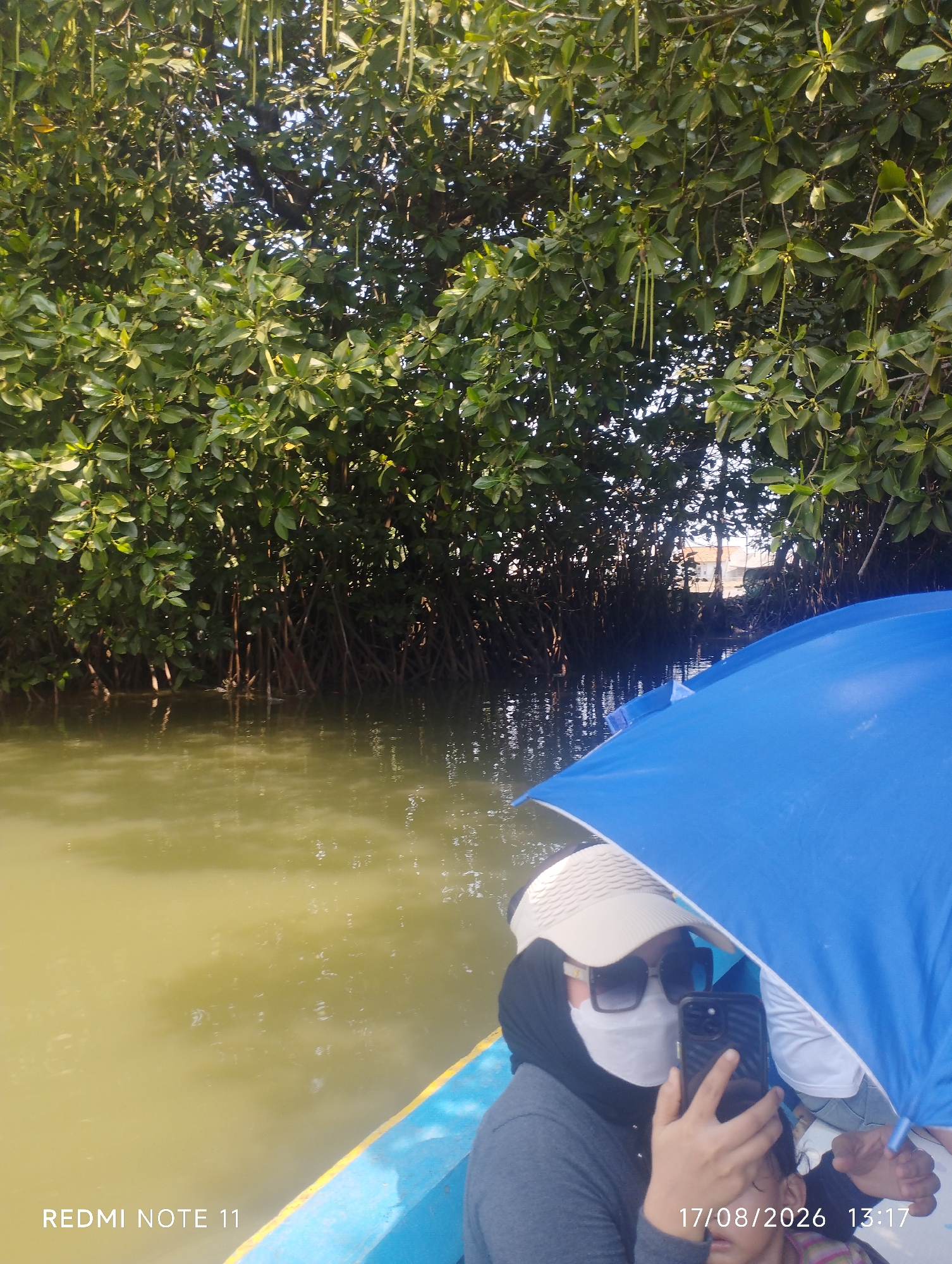 The image depicts a view from a boat on a body of water, likely a river or mangrove swamp, near Karawang, Indonesia, as indicated by the timestamp and phone model. A person wearing a hijab, sunglasses, and a white face mask is in the foreground, holding a smartphone and taking a selfie under a bright blue umbrella. Behind them, dense green mangrove trees with visible prop roots line the water's edge. The water is murky and reflects the surrounding foliage. The date and time "17/08/2026 13:17" are visible in the lower right corner. Another person, possibly a child, is partially visible behind the main subject. The overall scene suggests a tour or outing through a natural mangrove environment.