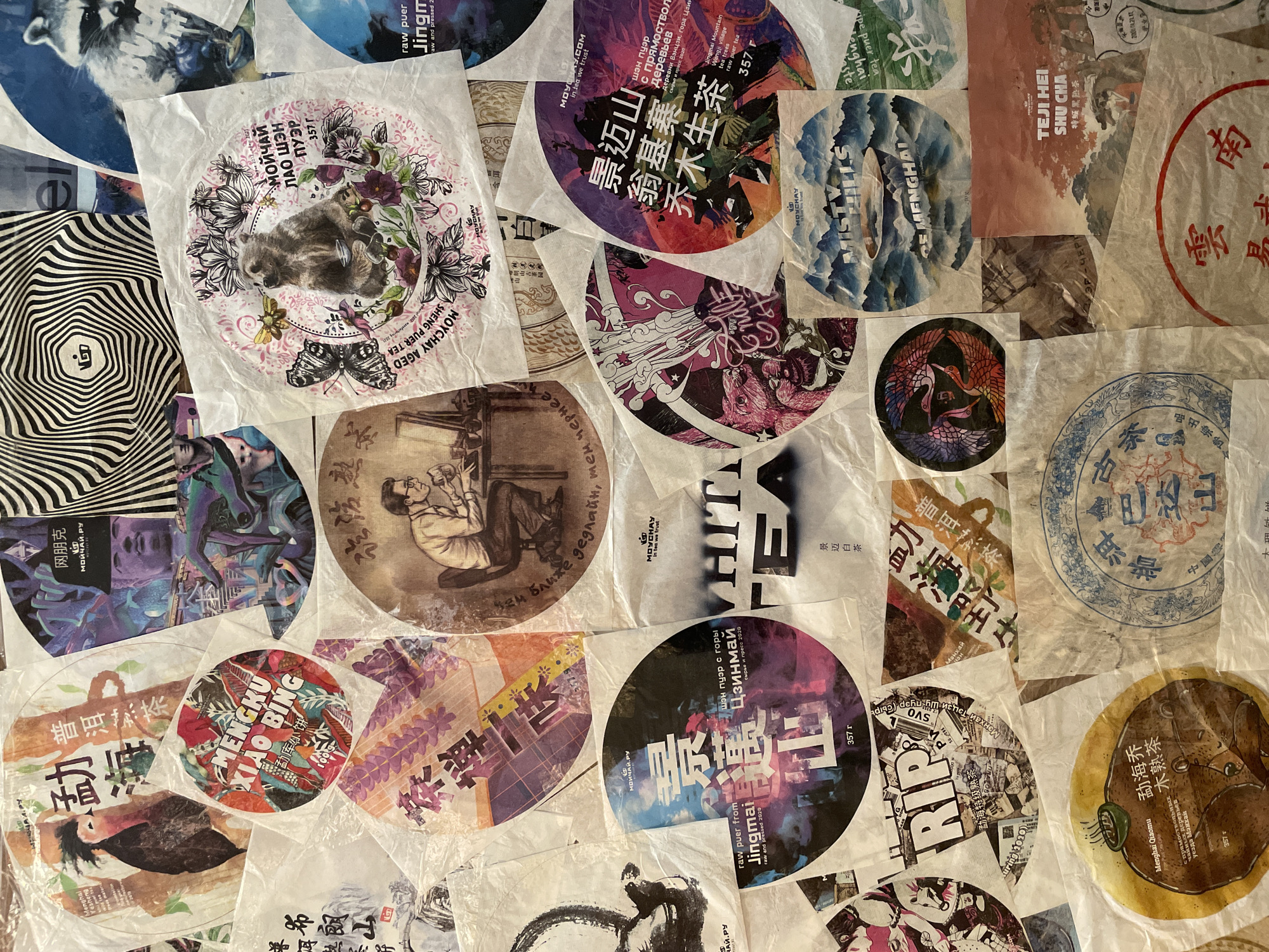 The image displays a close-up, overhead view of a collection of circular and semi-circular paper wrappers, likely for tea cakes, arranged in an overlapping, collage-like fashion on a light-colored surface. The thin, often translucent wrappers feature a wide variety of colorful and monochrome designs.
The subjects on the wrappers are diverse, including illustrations of animals such as a raccoon, a bear, cranes, and butterflies, alongside mythical creatures. Landscapes depicting mountains and trees, abstract patterns like swirling lines, and images of people, including a man reading and traditional figures, are also visible.
The predominant objects are these tea wrappers, showcasing intricate graphic design and typography. No specific activity is taking place; the image is a still life capturing an arrangement of collected items, possibly from a tea enthusiast or vendor.
Notable details include the presence of text in multiple languages: Chinese, Russian (Cyrillic), and English. Many wrappers identify the contents as various types of Puerh tea (Sheng Puer, Shu Cha, Aged Puer Tea), along with White Tea, and specify regions like "Jingmai Mountain," "Menghai," "Bulang Mountain," "Mengku," and "Yunnan." The brand "MOYCHAY" appears frequently. Weights such as "357 g" are common. Unique phrases like "the closer the deadline, the darker it gets" and "RIP" are also incorporated into the designs. The indoor lighting is even, suggesting an intentional display. This collection of wrappers is located in Tbilisi, Georgia.