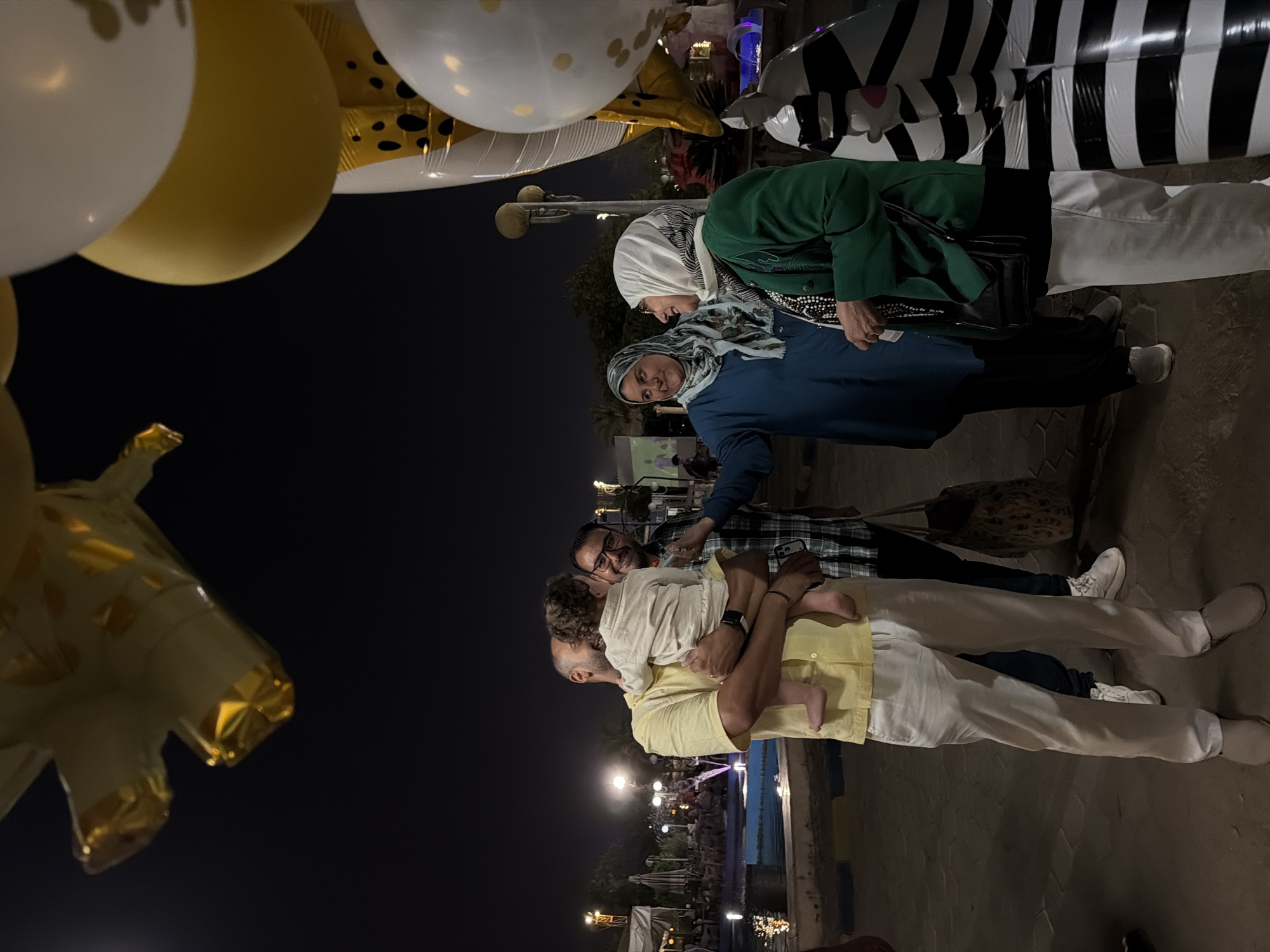 In the city of 6th of October, Egypt, at night, a group of people are gathered outdoors near a body of water. A man in a yellow shirt is holding a baby, facing a man in a plaid shirt who is smiling. To the right, two women wearing hijabs are talking; one in a blue dress and another in a green jacket. Balloons, including one shaped like a giraffe and round white and gold balloons, are visible in the upper part of the frame. A black and white striped inflatable animal, resembling a zebra, stands to the right of the women. The background shows some foliage, lights, and structures, suggesting an outdoor recreational area or a park. The lighting indicates it is nighttime.