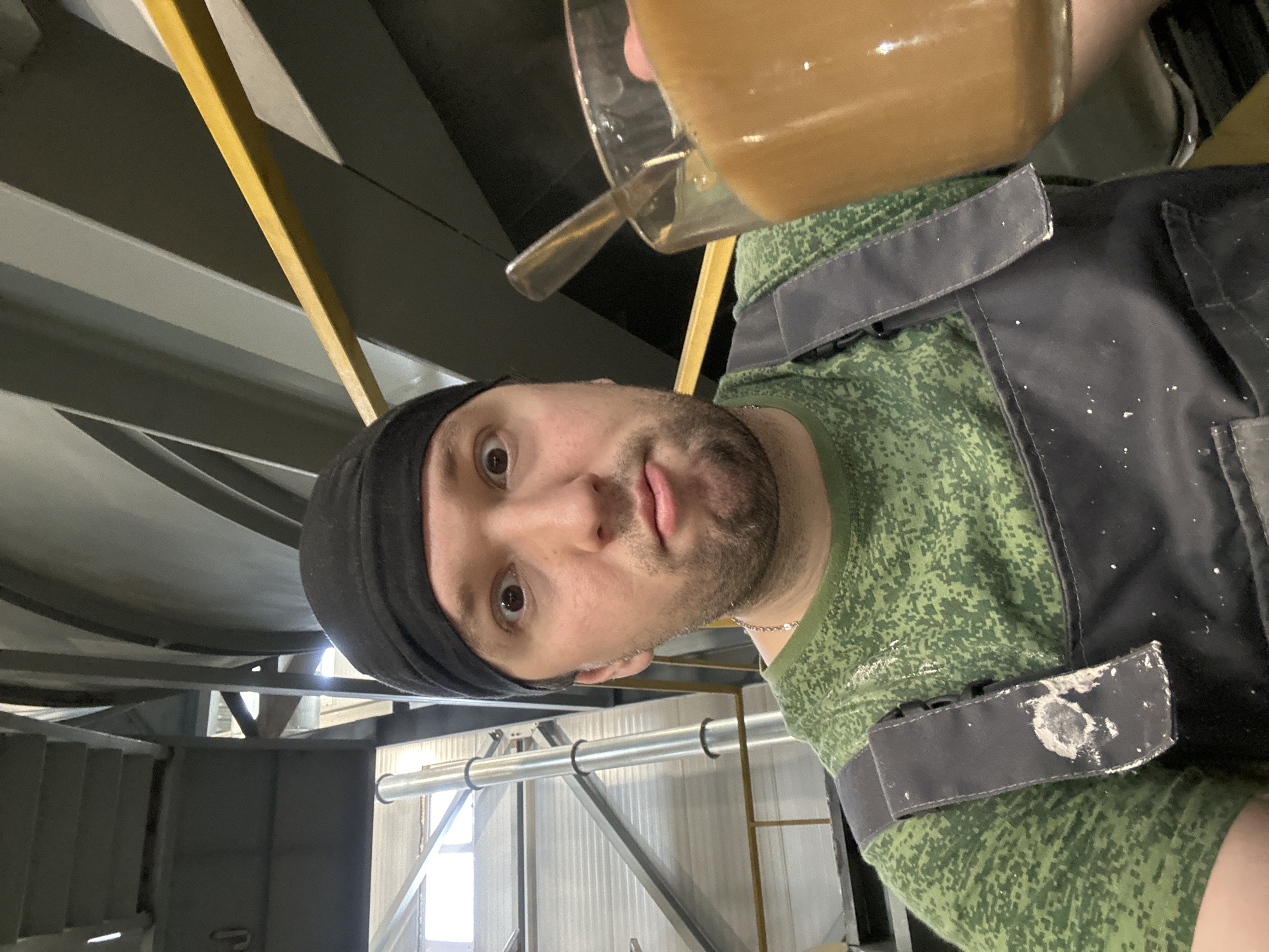 A man in a black head covering and green camouflage t-shirt with a black vest is holding a glass mug of brown liquid up to the camera. He appears to be making a face. The background appears to be an industrial setting with metal beams and a window in the distance. There is no indication the image was taken in Noviy, Russia.