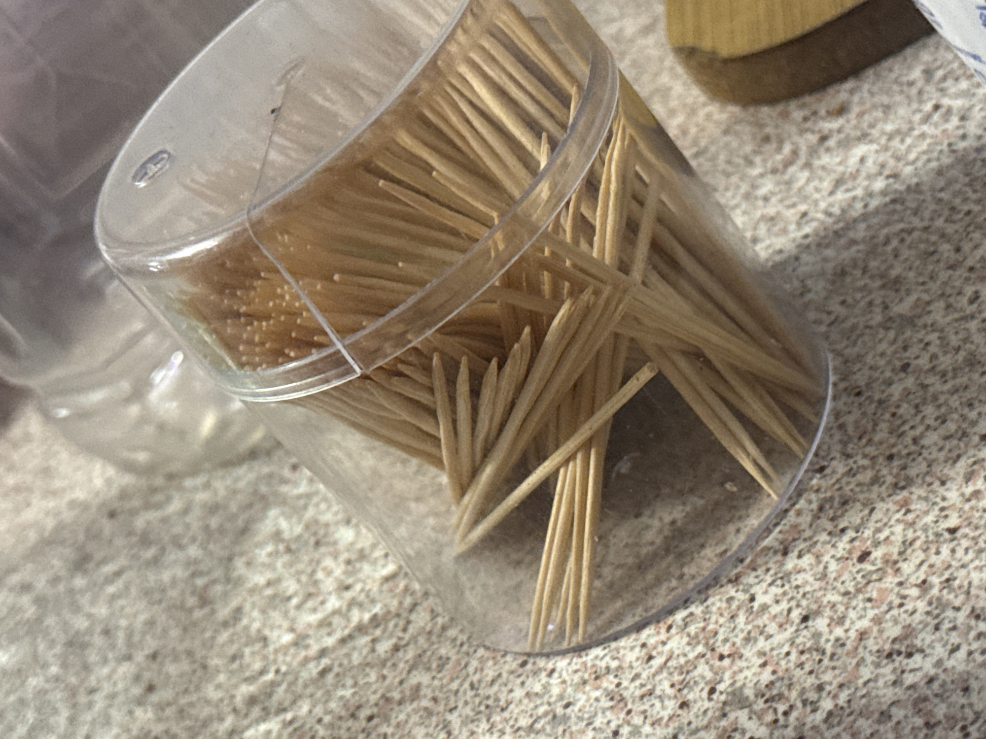 A clear, cylindrical plastic container filled with wooden toothpicks rests on a speckled, light-colored countertop. The container, positioned at an angle, is nearly full, with numerous toothpicks visible, some standing upright and others fallen or crisscrossed within. The lid of the container is also clear plastic and features a small, oval-shaped opening or dispenser. To the right, a dark, small speck is visible on the inner surface of the lid. The countertop has a mottled pattern of light gray, beige, and darker flecks. In the blurry background, indistinct shapes of other translucent containers and a wooden block-like object are partially visible. Shadows are cast on the countertop, suggesting illumination from an overhead or angled light source. No individuals are present, and no specific actions are taking place. The scene provides no discernible details to indicate a location within the city of Varna, Bulgaria.