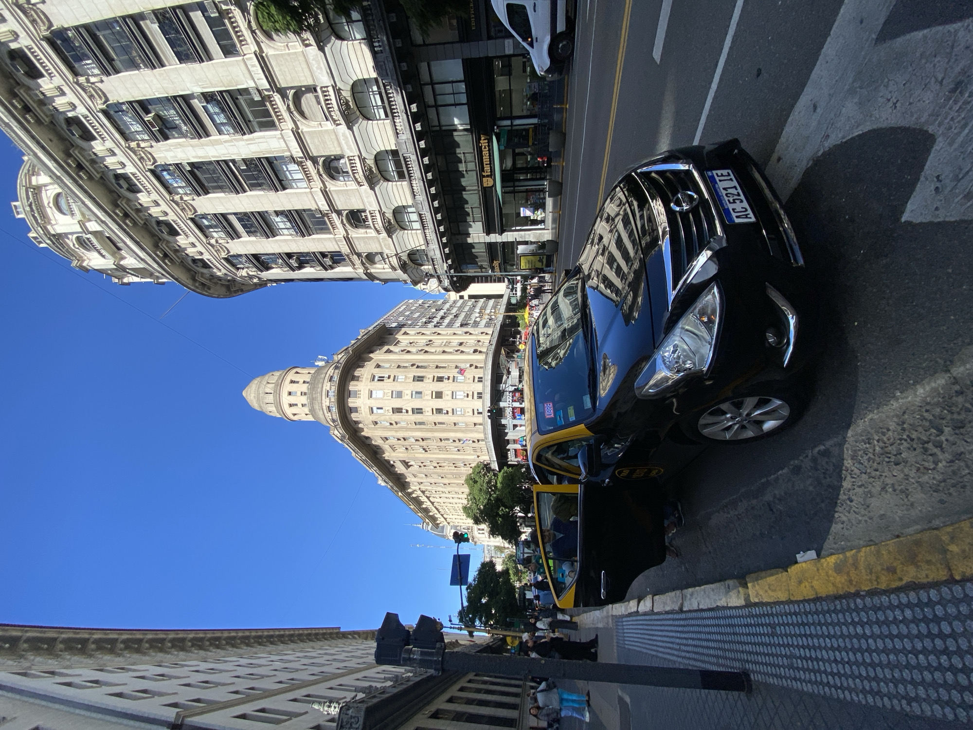 The image shows a street scene in Buenos Aires, Argentina. A black taxi with the license plate "AC 521 IE" is parked on the right side of the street, with its passenger door open. The sidewalk has a textured tactile paving surface. Several people are on the sidewalk. The street has yellow lane markings. Buildings with ornate facades line both sides of the street, typical of the architecture in Buenos Aires. One building has a rounded corner topped with a dome. A "farmacity" store is visible in one of the buildings.