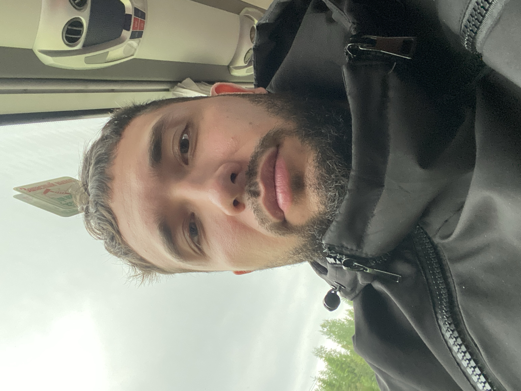 A close-up selfie of a man with a beard. He is indoors, possibly on public transportation, based on the interior ceiling fixtures visible in the background. He is wearing a black jacket. Beyond the window, there are trees and an overcast sky. The location in the city of Bereznyaki, Russia is not discernable in the image.