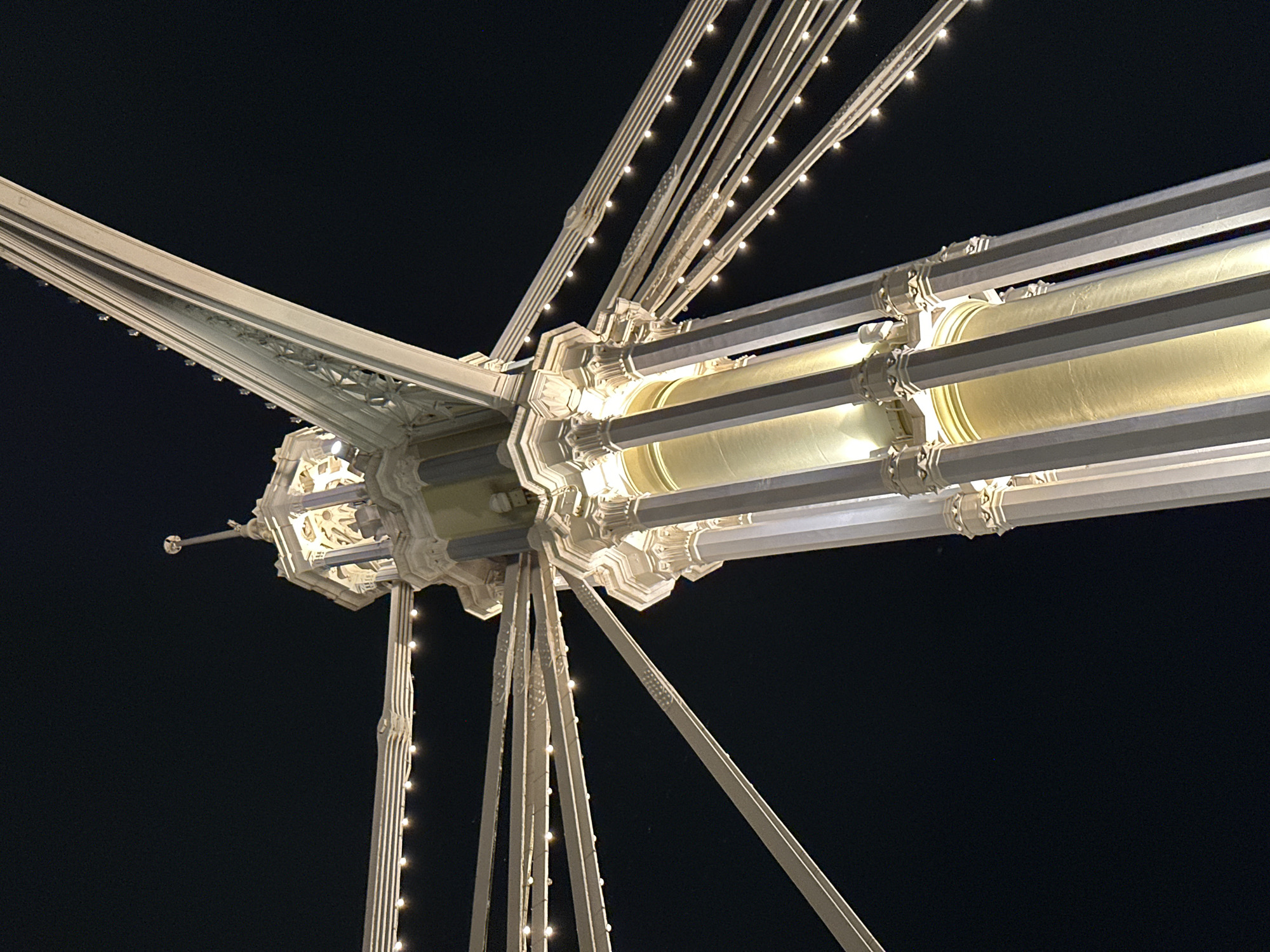 The image presents a detailed, low-angle view of an illuminated architectural structure set against a deep black night sky. The primary subject is a complex arrangement of white or light-colored metal beams and cables converging at a central, ornate hub. This central connector features intricate decorative elements, including fluted columns, corbels, and faceted sections, suggestive of a classical or Victorian architectural style.
Numerous thin cables, strung with small, bright white lights, radiate outwards from the central hub and extend along the main beams, creating a dazzling effect against the darkness. Larger, cylindrical elements, possibly light fixtures or decorative casings, are integrated into the horizontal beams. These emit a warm, golden glow, illuminating sections of the structure and contrasting with the cooler white light of the smaller bulbs.
The image captures the structure in static repose; no activity or event is taking place, emphasizing its intricate design. The perfectly dark background indicates the photograph was taken at night. The artificial illumination highlights the grandeur and elaborate detailing of the structure. Given the context of London, United Kingdom, these architectural and lighting characteristics strongly resemble details found on landmark structures such as Tower Bridge. There is no visible text in the image.