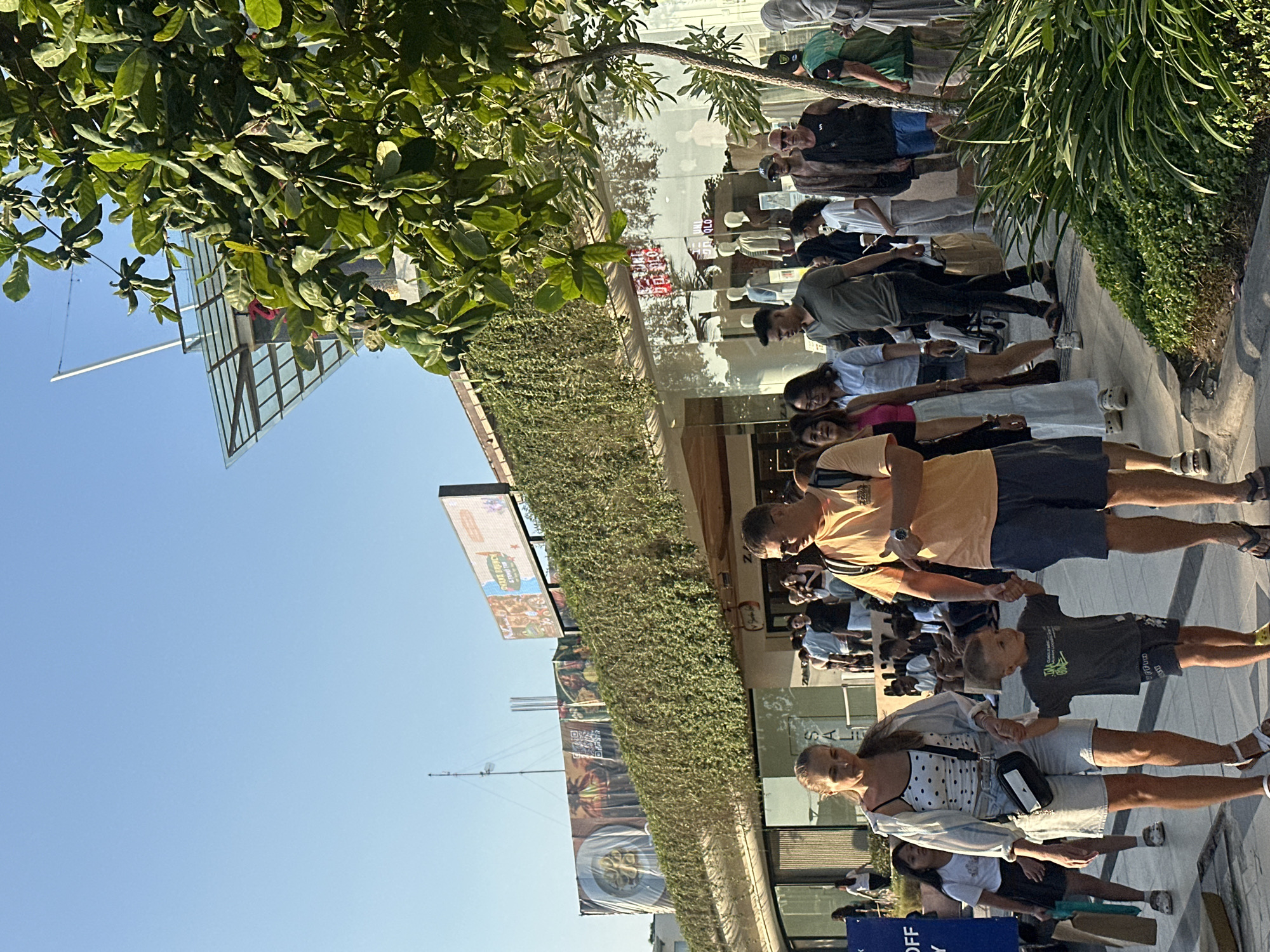In Kuta, Indonesia, a family walks on a sunlit pedestrian pathway. A woman in a white dotted top and denim shorts holds hands with a young boy in a grey t-shirt and dark shorts, who is walking ahead of her. A man in an orange t-shirt and blue shorts walks to the right of the woman and boy, also holding the boy's hand. Behind them, a crowd of people walks in the same direction, with shops visible behind them, characterized by large glass windows and an overhang covered in greenery. The sky is clear and blue.