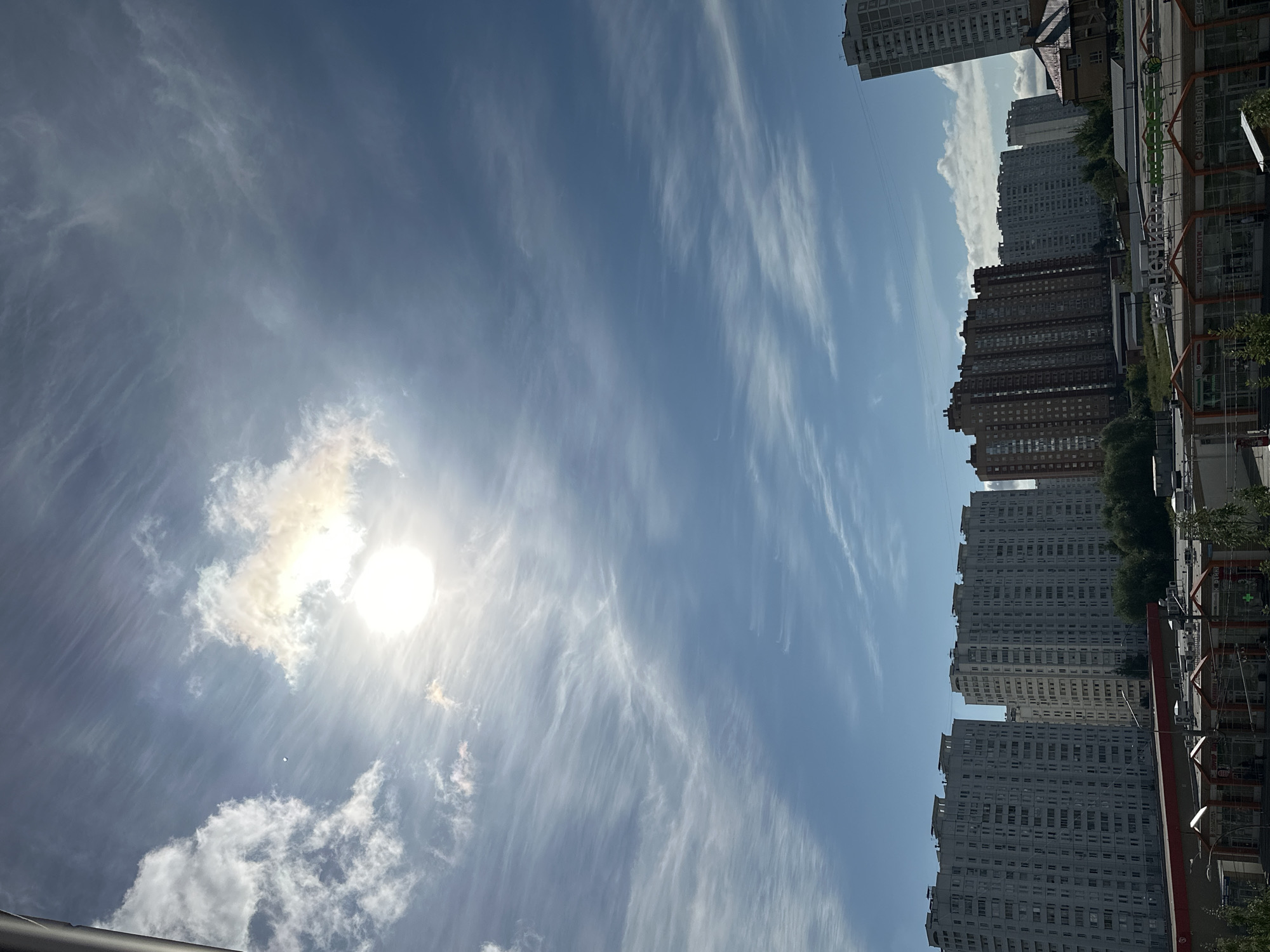 The image depicts a view of Podolsk, Russia, featuring a skyline dominated by residential buildings under a bright, partially cloudy sky. In the foreground, a row of single-story commercial buildings with red awnings is visible, suggesting a shopping area. Behind these, several multi-story apartment complexes of varying architectural styles and colors punctuate the horizon. The sky is a prominent feature, with the sun shining brightly through wispy cirrus clouds, creating a dynamic and somewhat hazy atmosphere. The overall scene suggests a typical urban environment in Podolsk on a sunny day.