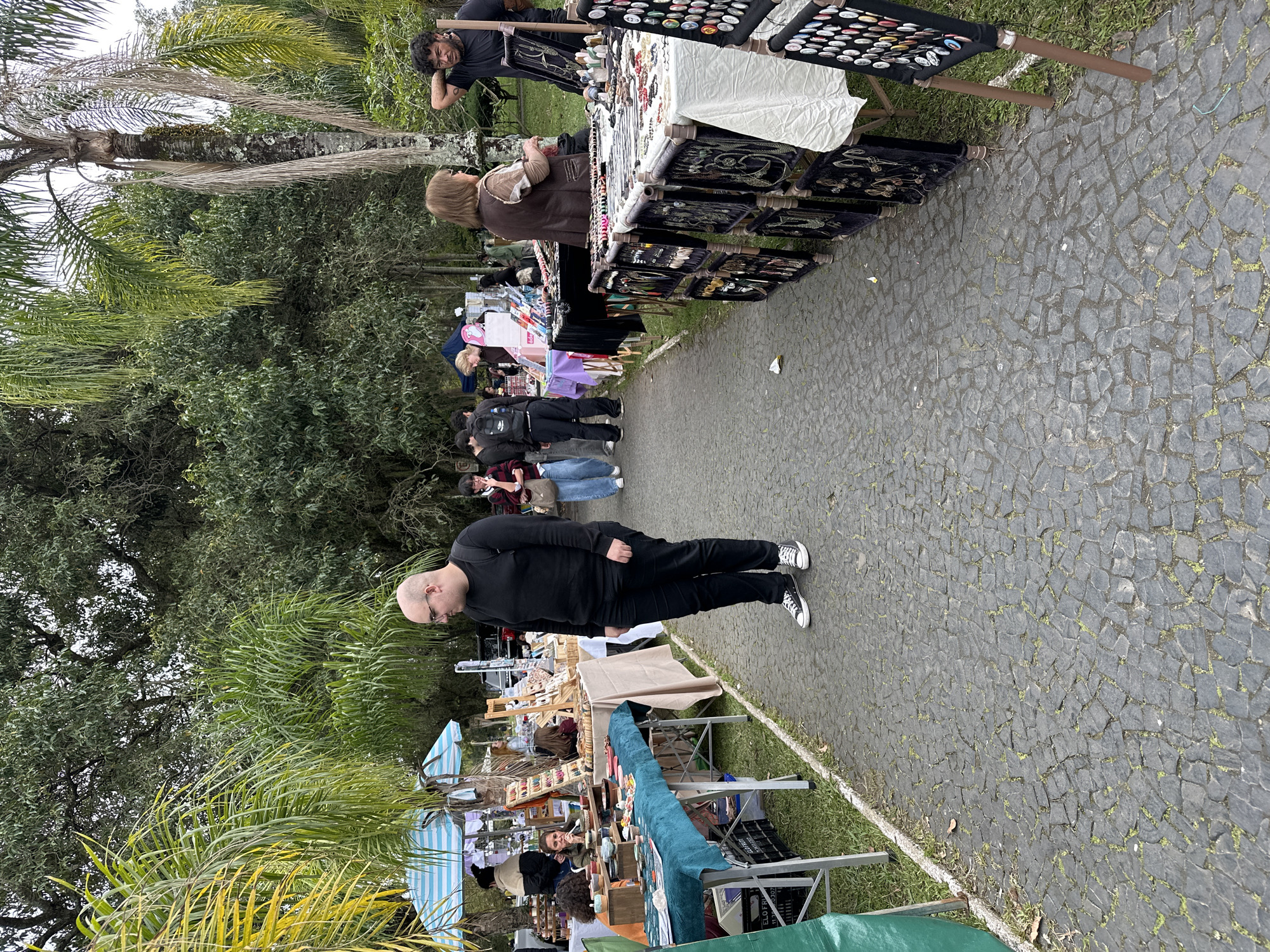 This image depicts an outdoor market scene, likely in Florianópolis, Brazil, with cobblestone paving and lush greenery. Several people are browsing or manning stalls displaying various items, including jewelry, crafts, and potentially clothing. The foreground features a bald man wearing a black long-sleeved shirt and black pants, looking towards the stalls. Behind him, to the left, are stalls with items displayed on tables covered with colorful cloths. Further into the scene, more stalls are visible, laden with jewelry and trinkets, with several individuals engaged in activities related to shopping or selling. Palm trees and dense foliage frame the market, suggesting a tropical or subtropical environment. The overall atmosphere appears to be casual and bustling, characteristic of a local market.