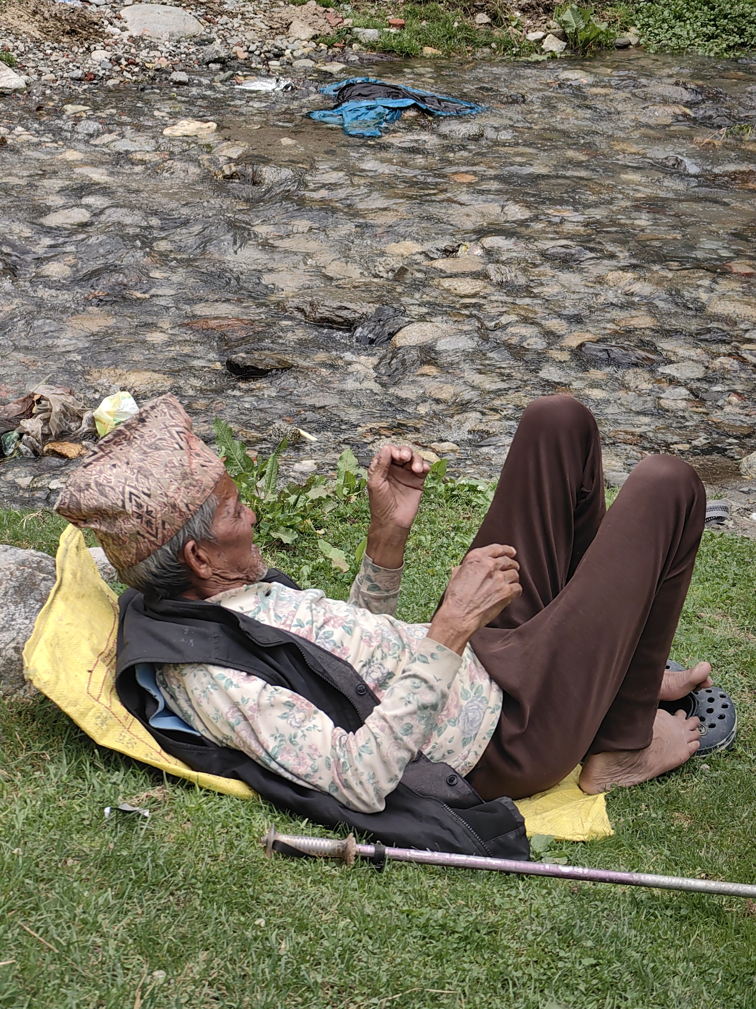 An elderly individual with weathered skin, wearing a patterned cap and a floral-print shirt, rests on a yellow mat on a grassy area next to a rocky stream in Vashisht, India. The individual is lying on their back with their legs extended, one foot shod in a black clog. They are wearing brown trousers and a dark vest over their shirt. A trekking pole lies on the grass in front of them, and their hands are resting on their lap. The stream bed is filled with stones and clear water, with some debris, including blue plastic, visible in the water. Green vegetation grows along the bank of the stream. The overall scene depicts a moment of rest in a natural, outdoor setting.