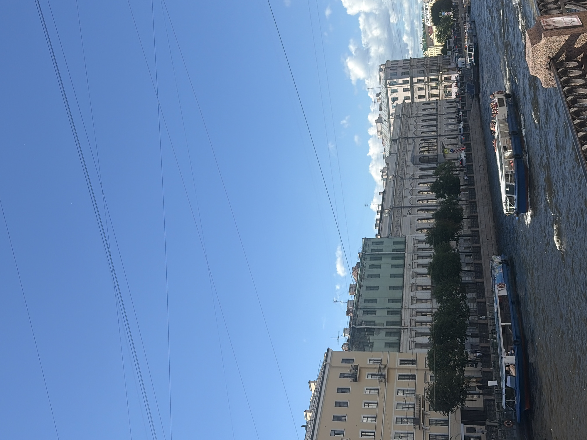 This image depicts a canal scene in Saint Petersburg, Russia, on a bright, sunny day with scattered clouds. The perspective is from above, looking down at the canal and the buildings lining its edge. Several boats are visible on the water, including a blue and white tour boat filled with passengers. The architecture of the buildings suggests a historical European city, with ornate facades and multiple stories. Trees line the canal in front of the buildings, providing a touch of greenery. Numerous overhead wires crisscross the sky, a common sight in urban environments. The water of the canal is rippled, indicating movement, likely from the boats. The overall impression is a lively, urban waterway on a pleasant day.