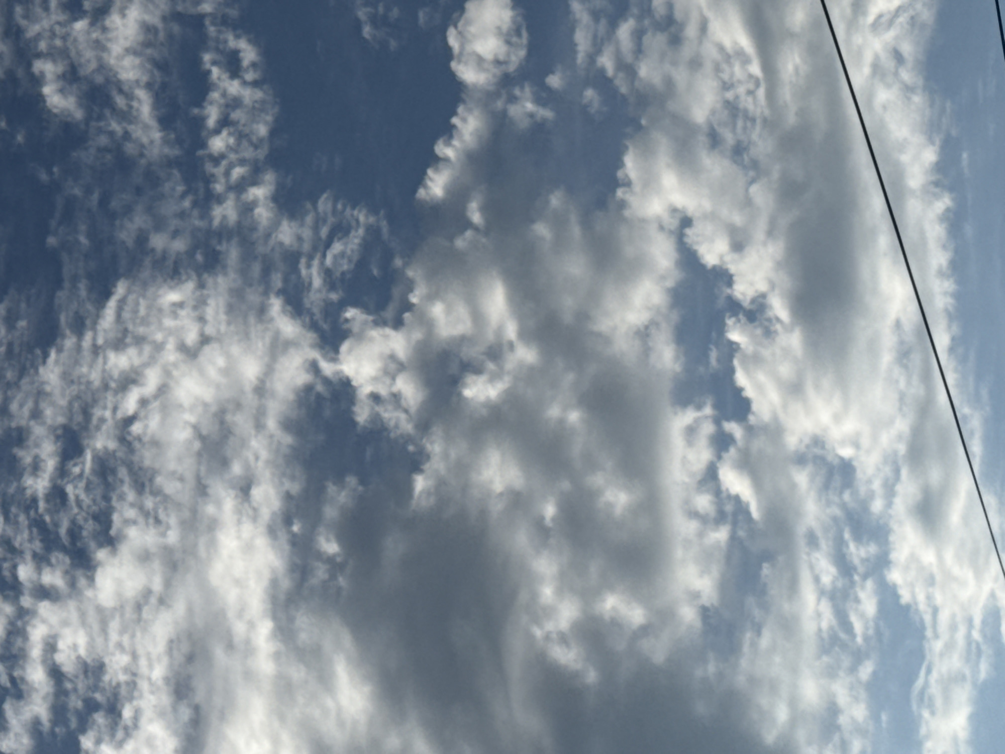 This media file depicts a cloudy sky with a dark wire running diagonally from the top right to the bottom left. The sky is a mix of deep blue and lighter shades, with numerous white and gray clouds scattered across it. Some clouds appear thin and wispy, while others are thicker and more opaque, suggesting varying levels of moisture and atmospheric conditions. The overall impression is of a dynamic sky, possibly indicative of changing weather. The presence of the wire suggests an outdoor setting, likely with human infrastructure nearby, though no specific structures or landscape features are visible. The lighting suggests it is daytime, likely late afternoon or early evening given the slight dimness and the dramatic cloud formations. No text is visible in the image.