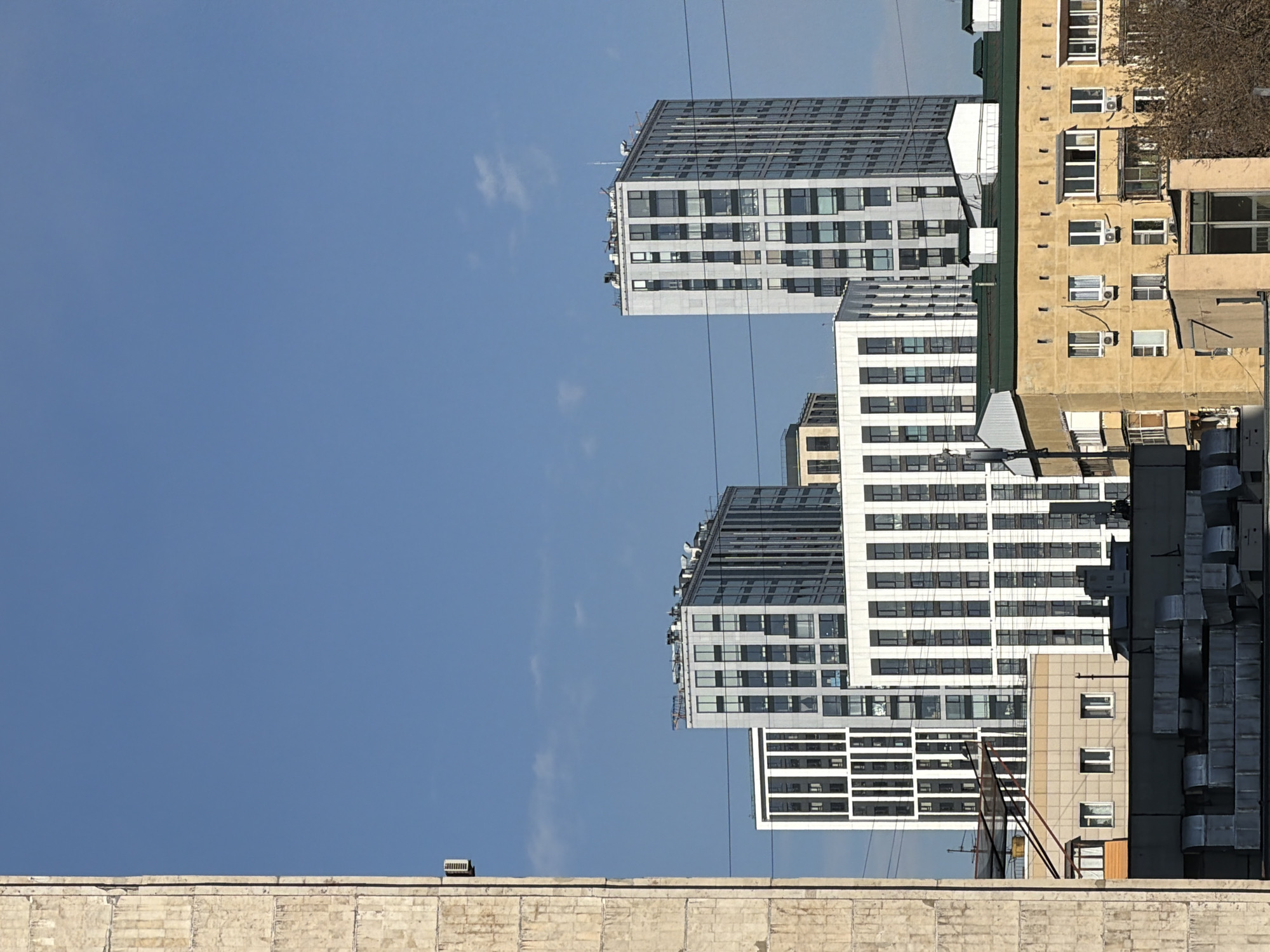The image presents a view of buildings and infrastructure against a blue sky, likely in an urban setting. In the foreground, there is a low brick wall or rooftop. Behind it, there are several buildings of varying heights and architectural styles. The structures include modern apartment blocks with grids of windows and older residential buildings. A network of wires runs in front of the buildings, suggesting the presence of power lines or communication cables. The angle of the photo suggests it might have been taken from a slightly lower vantage point, looking upwards. Given the architecture and appearance of the environment, it is reasonable to speculate that the location is Moscow, Russia, with a mix of Soviet-era and contemporary buildings.
