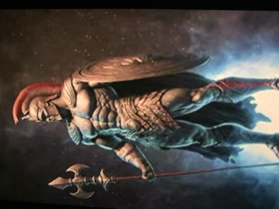 The image displays a full-body, stylized digital rendering of a highly muscular, armored warrior standing in a static pose. The figure, facing right with its head turned to the left, wears a bronze-toned helmet featuring a prominent red plumed crest. Its armor consists of intricate shoulder pads, a segmented cuirass emphasizing defined abdominal musculature, a scaled skirt, and ornate leg greaves and arm bracers. A dark cape drapes over the figure's left shoulder. In its right hand, it grips a tall, ornate spear with a complex, multi-bladed head and a reddish shaft. A large, circular shield with concentric ring patterns is held on its left arm. The background is dark and nebulous, speckled with faint luminous particles, suggesting an ethereal or cosmic environment. A strong, electric blue light emanates from beneath and around the warrior's lower half, casting dramatic upward illumination.
Location: Jos, Nigeria.