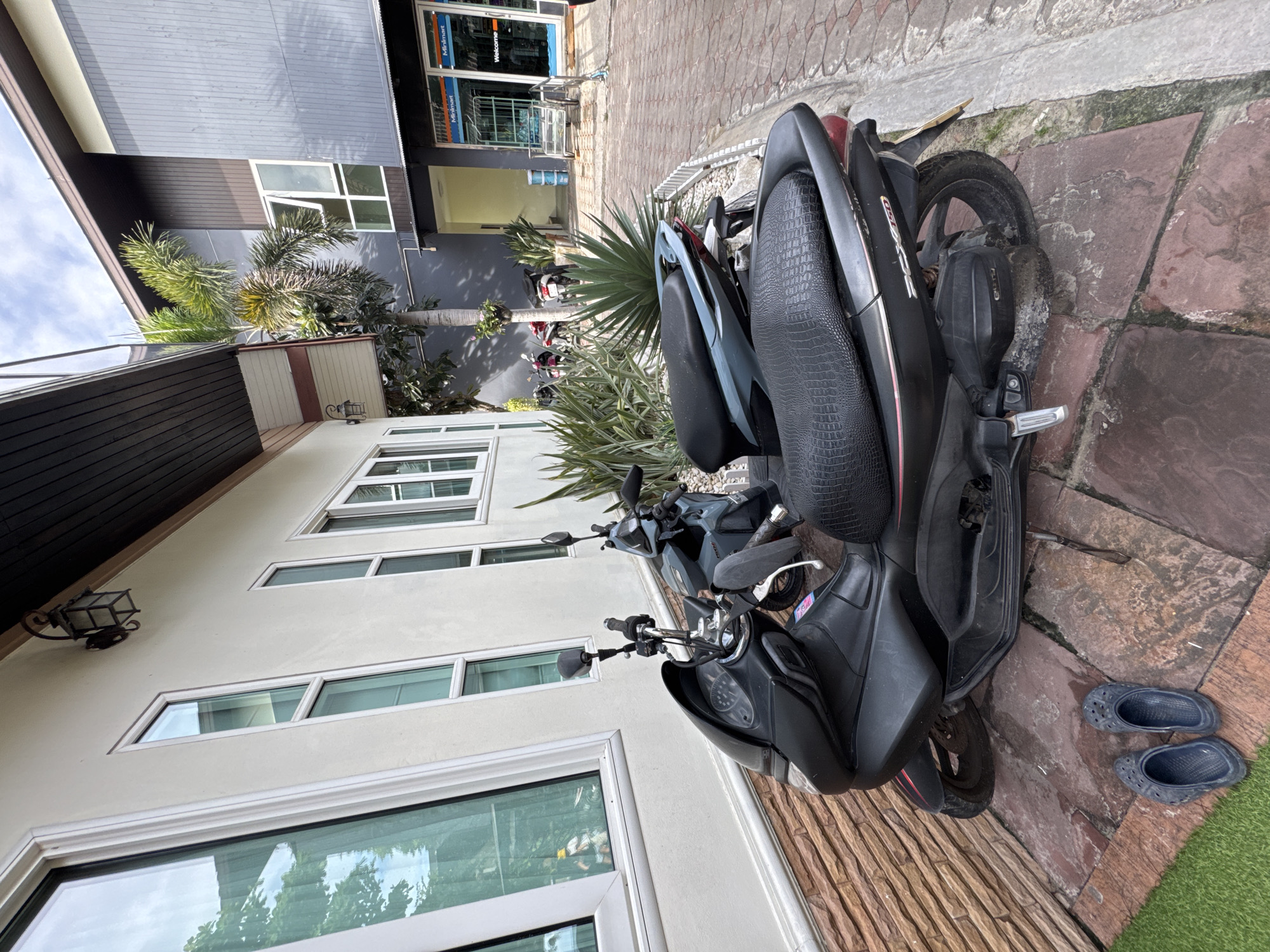 The image depicts a street scene in Ao Phai, Thailand, with two parked scooters in the foreground. The scooter on the left is black with a textured seat and red accents, positioned on a stone patio. A pair of dark blue clogs are placed on the patio to the left of this scooter. Behind it, a second scooter, light blue and grey, is also parked. The scooters are situated next to a building with a light-colored facade, large windows reflecting trees and sky, and a dark brown overhang. A black lantern is mounted on the wall above the windows. In the background, a multi-story building with grey and dark brown siding is visible, along with a palm tree and lush green foliage. Further back, a minimart with glass doors and signage is present. The ground is paved with reddish-brown rectangular stones and some green artificial turf. The sky above is partly cloudy.