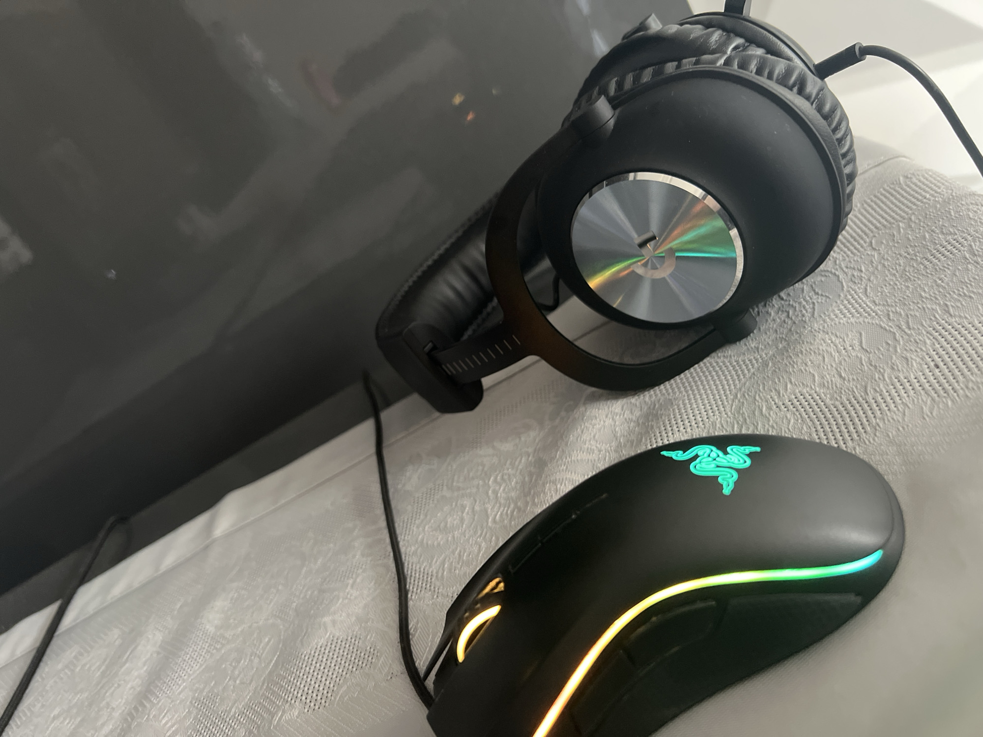 The image depicts a black Logitech G Pro X gaming headset and a black Razer gaming mouse with illuminated RGB lighting on a white patterned surface. The headset is positioned near the upper right of the image, resting slightly on its side. A cable extends from the headset. The mouse is located below and slightly to the left of the headset. The image is angled, with a grey, possibly reflective, surface appearing in the background on the left side. The location in Burgas, Bulgaria is not discernible from the image.