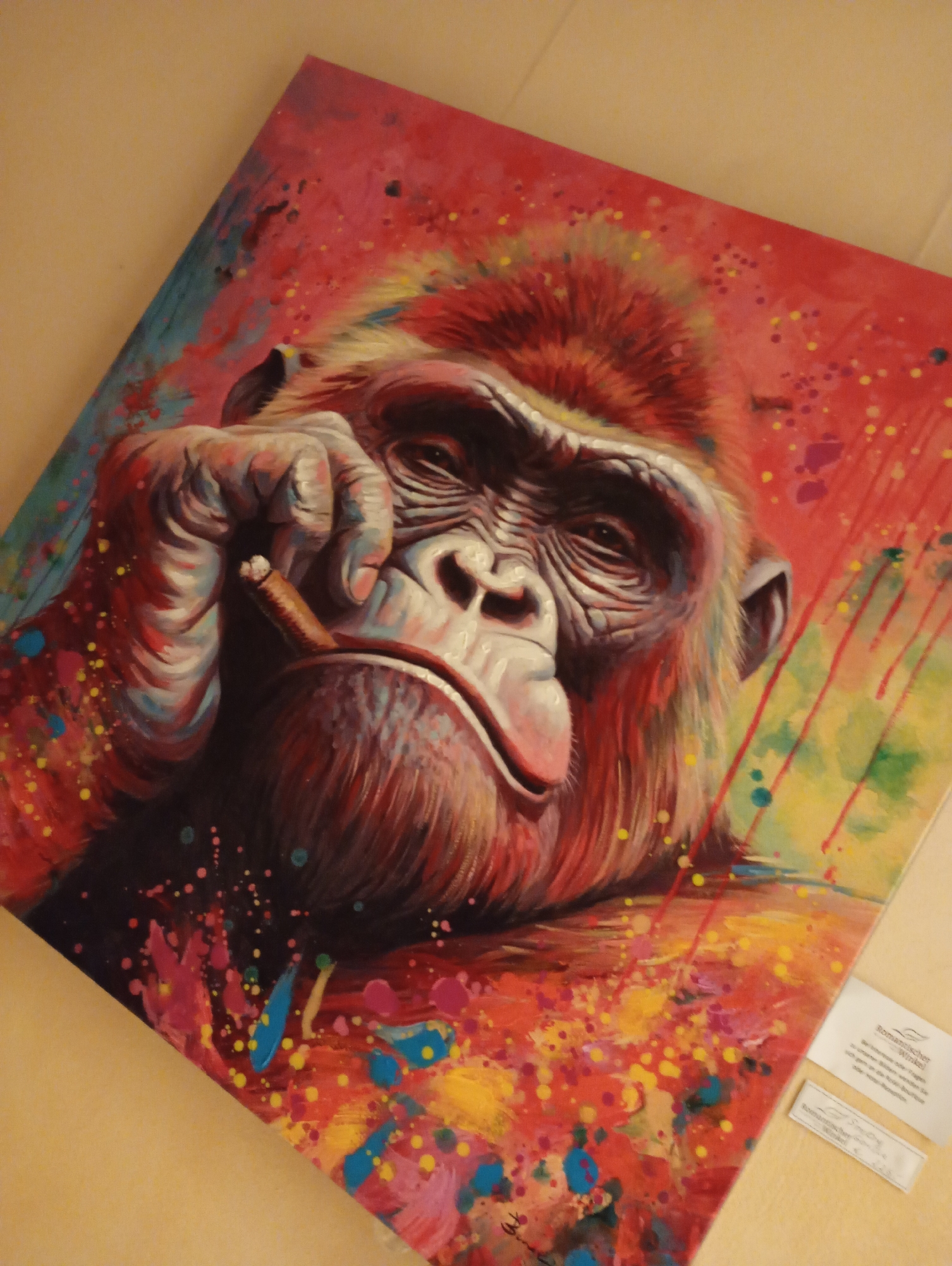 The image displays a close-up, angled shot of a vibrant, abstract painting featuring a chimpanzee. The chimpanzee is depicted in a portrait style, holding a lit cigar to its mouth with its left hand. Its expression appears contemplative or world-weary, with visible wrinkles around its eyes and muzzle. The artwork is characterized by a bold color palette, dominated by reds, oranges, yellows, and splashes of blue and pink, creating a dynamic and energetic background. The painting is positioned on a light-colored surface, possibly a table or floor, and is angled to show a label attached to its lower right corner. The label appears to be a product or artist information tag, written in German. The overall impression is one of a contemporary art piece, likely displayed or stored in a residential or studio setting. The specific location of Bad Sachsa, Germany is not visually evident within the image itself, but the presence of German text on the label suggests a connection to that region.