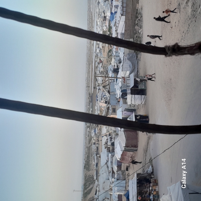 A view of a crowded refugee camp is captured. The image is taken from a slightly elevated position, looking down upon a vast expanse of makeshift shelters. Individuals are seen moving around, highlighting the densely populated nature of the camp. Two thick, dark beams frame the view, possibly parts of the structure where the photo was taken. The sky is light, suggesting either dawn or dusk.