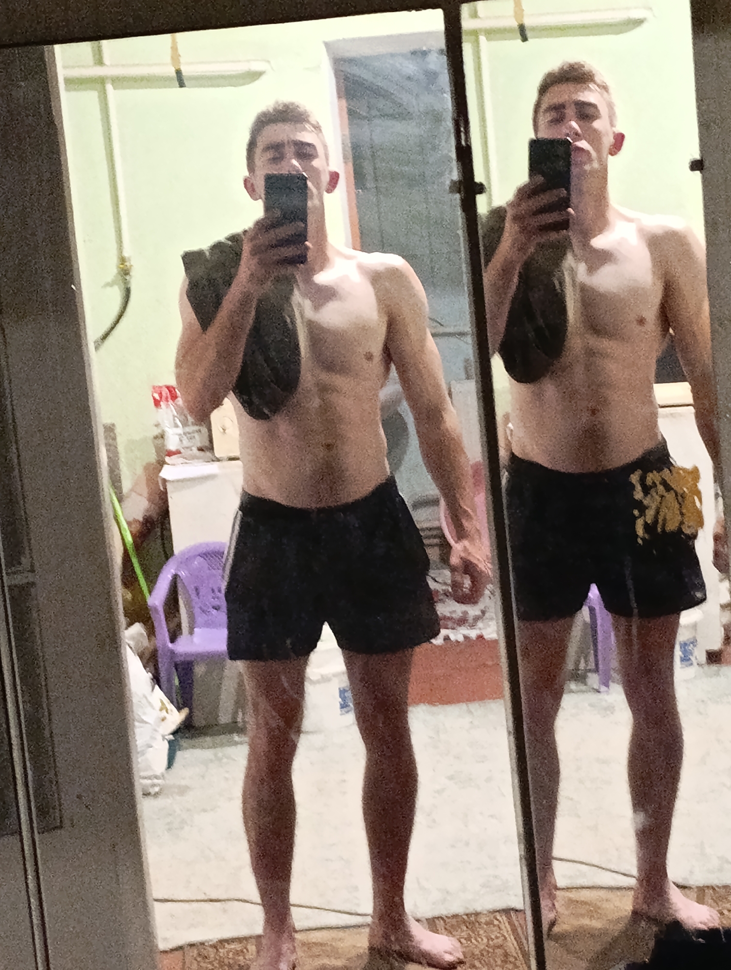 The image shows a young man taking a mirror selfie in what appears to be a bathroom. He is shirtless and wearing black athletic shorts. A dark-colored garment is draped over his shoulder. The background is cluttered with various items, including bottles, a purple chair, and what seems to be cleaning supplies. The walls are painted a light green. The overall impression is of a casual, indoor setting. There is no discernible information within the image to suggest a location in Fastiv, Ukraine.