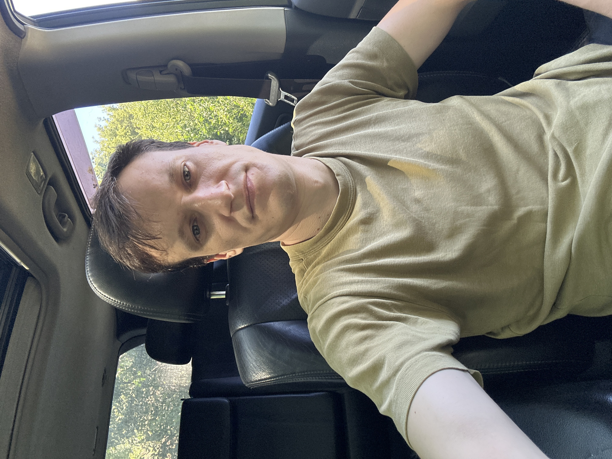 A young Caucasian man is pictured inside a car. He is wearing a light olive green t-shirt and is seated in the driver's seat, facing the camera. The car's interior is visible, with black leather seats and a sunroof. Outside the car, lush green trees can be seen. The scene appears to be in Shatava, Ukraine, given the context provided. The lighting suggests it is daytime, possibly with sunlight filtering through the trees and windows. The man's expression is neutral. The image is taken from a close-up perspective, focusing on the man and the immediate interior of the car.