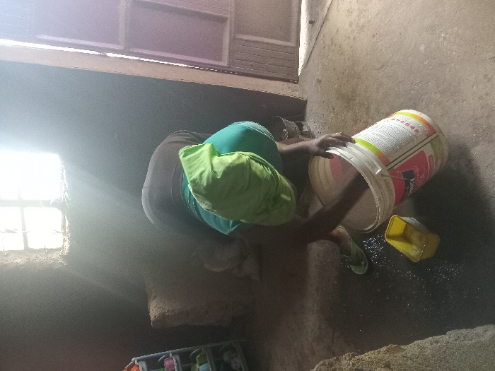 An indoor scene in Manda-Shivanga ward, Kenya, depicts an individual bent forward, actively engaged with a large white bucket. The person wears a dark upper garment, a light green fabric over their back/shoulders, and light green flip-flops on their feet. Their hands are submerged inside the white bucket, which features a multi-colored label including the word "APEXA." Adjacent to the bucket on the dark, damp floor is a yellow rectangular container holding a clear liquid. Splashed liquid is visible on the floor around these items. A bright, gridded window high on the rear wall illuminates the space with strong natural light, casting visible rays through the otherwise dimly lit room. The walls are dark and textured. To the far left, a light-colored, multi-tiered shelving unit holds various small, colorful plastic items. A light-colored door with vertical paneling is partially visible on the right side of the frame. The floor appears to be concrete or compacted earth.