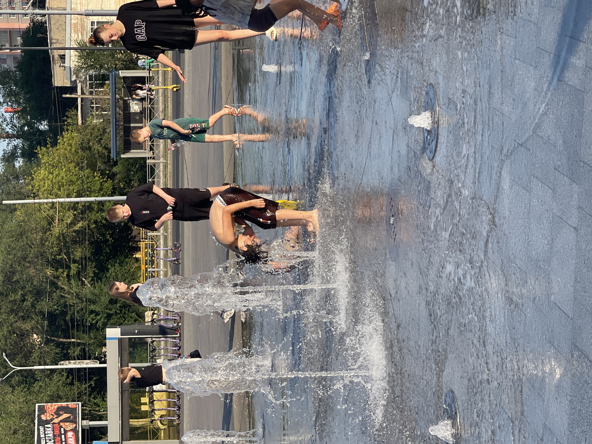 A group of children are playing in a public fountain in Orenburg, Russia. The fountain is located on a paved area with multiple spray nozzles. The children are dressed in casual summer clothing and are splashing in the water. One child is bending down, appearing to be playing with the water. In the background, there are trees, buildings, and what appear to be electric scooters parked in a designated area. The sun is shining, casting shadows on the wet pavement.