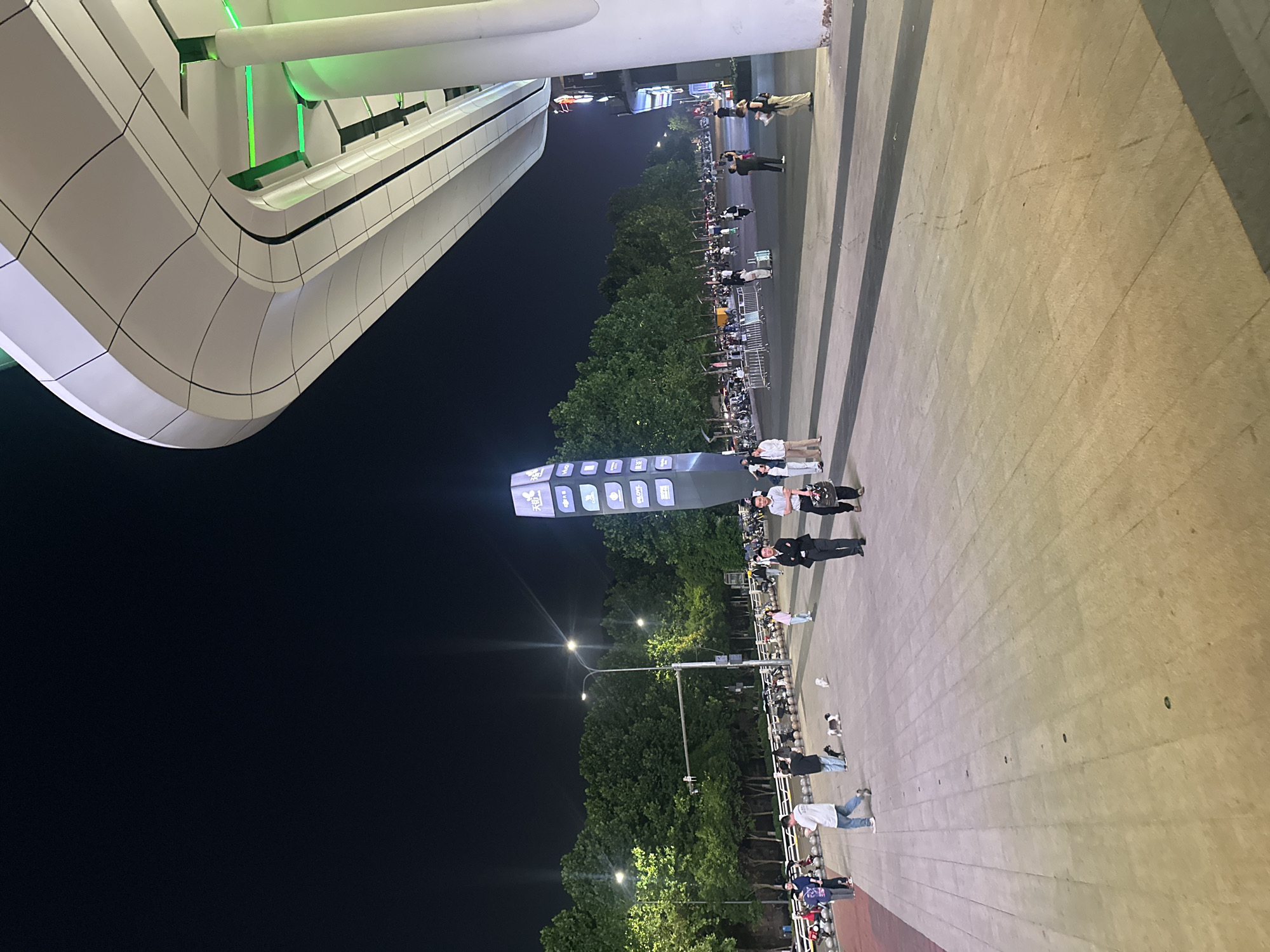 The image depicts an outdoor scene at night, likely in a public urban area. In the foreground, a paved walkway with a geometric pattern is visible, leading towards the right side of the frame. The walkway appears to be made of large rectangular tiles.
To the right, a modern architectural structure with a curved, white facade dominates the upper portion of the image. This structure features integrated green lighting, highlighting its sleek design. White cylindrical pillars support the overhang.
In the mid-ground, a diverse group of people are walking on the paved area. Some individuals are dressed casually, while others are in more formal attire. A tall, illuminated digital display tower with various logos and text is prominently situated to the left of the people. Trees line the background, forming a dark green canopy against the night sky. Streetlights with visible beams illuminate the area, casting a warm glow.
Further in the background, parked motorcycles and scooters are visible, suggesting a transportation hub or a busy area. Fences and barriers are also present, delineating spaces. The overall atmosphere appears to be lively, with people engaged in various activities, such as walking and socializing. The scene is set under a dark night sky, illuminated by artificial lights from the structures and street lamps. The location is identified as the Chongqing High-tech Zone, China.