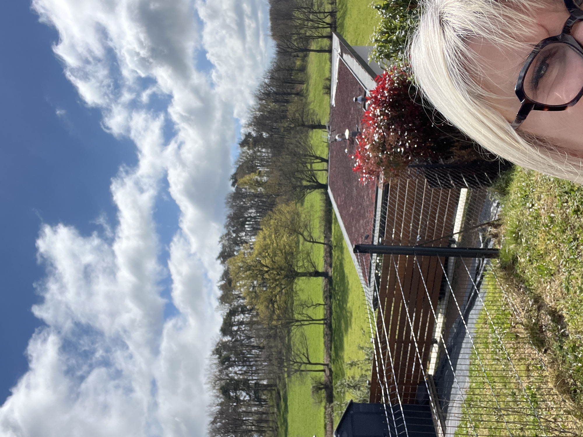 A sunny outdoor scene features a light-skinned person with blonde hair and glasses in the upper right corner. To the left is a view of a green landscape with a line of trees bordering a bright green field, punctuated by several other trees. A building with a red-brown roof is visible, and a bush with red and white flowers is next to the woman. A metal fence is running along the bottom right of the image. The sky is a bright blue with fluffy white clouds. The location is Chambœuf, France.